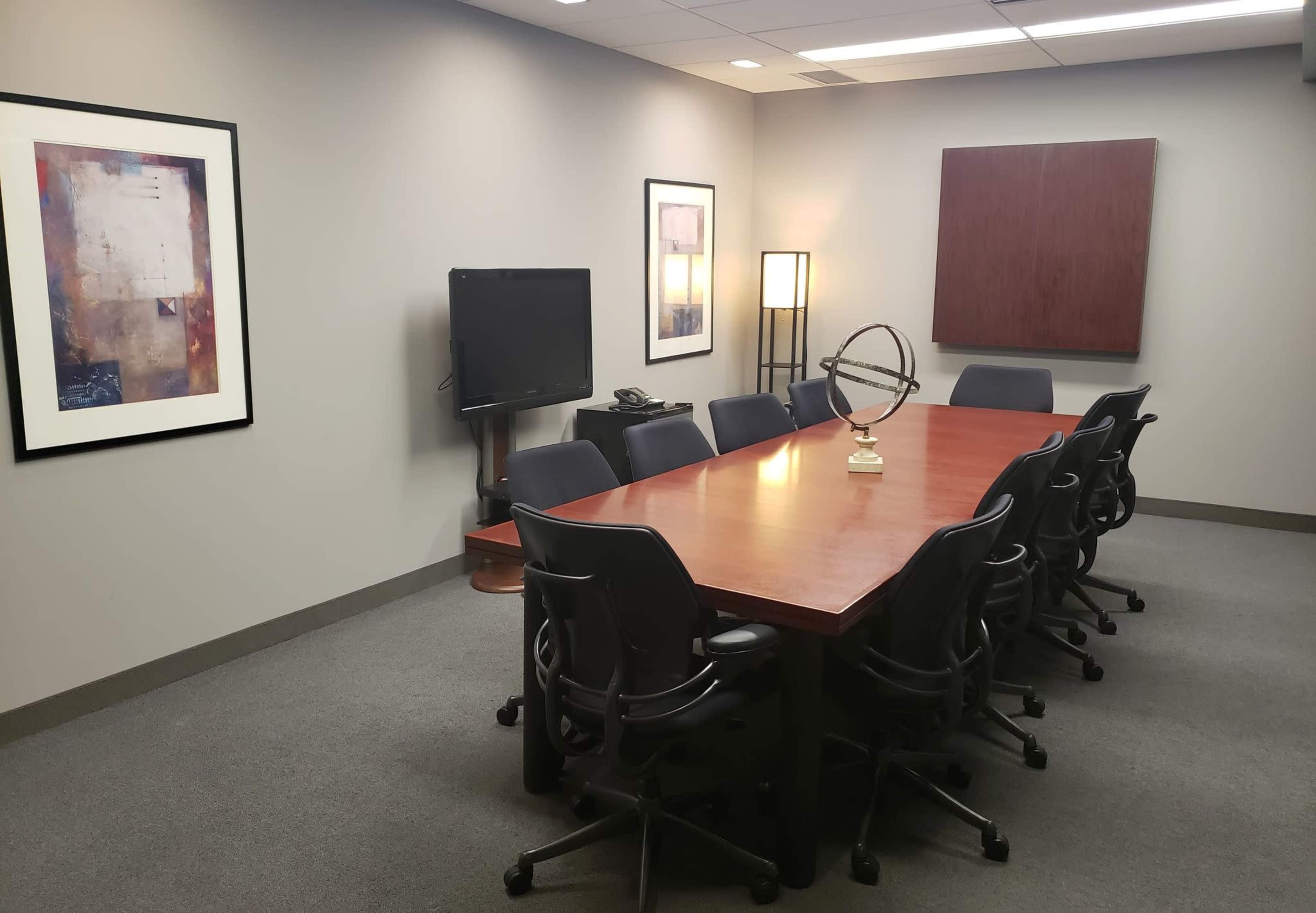 A conference room with a large wooden table surrounded by chairs, a television on the wall, and framed artwork.