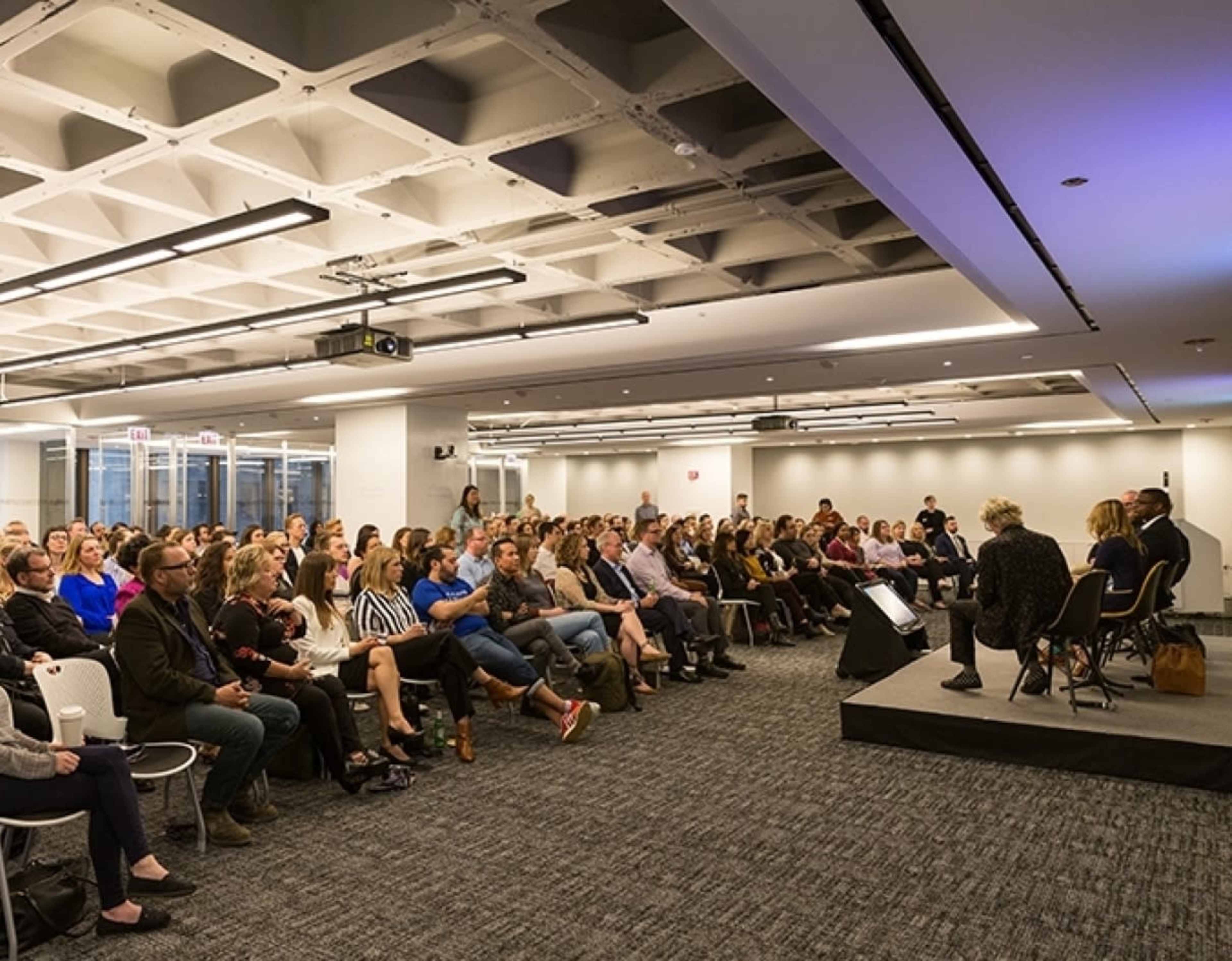 A large audience is seated in a modern conference room, watching a panel discussion on stage.