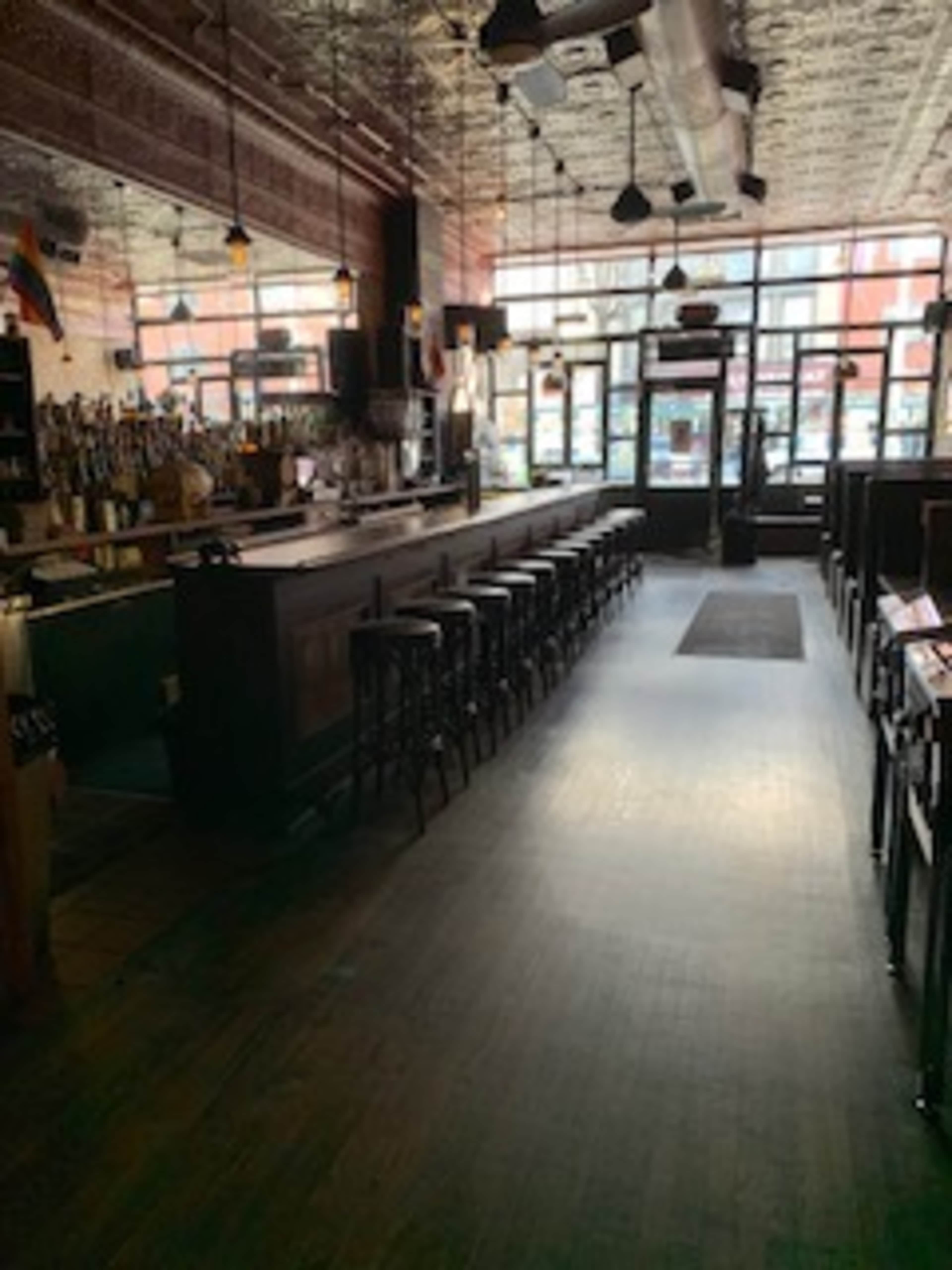 The image shows a dimly lit bar with a long counter, high stools, and large windows letting in natural light.