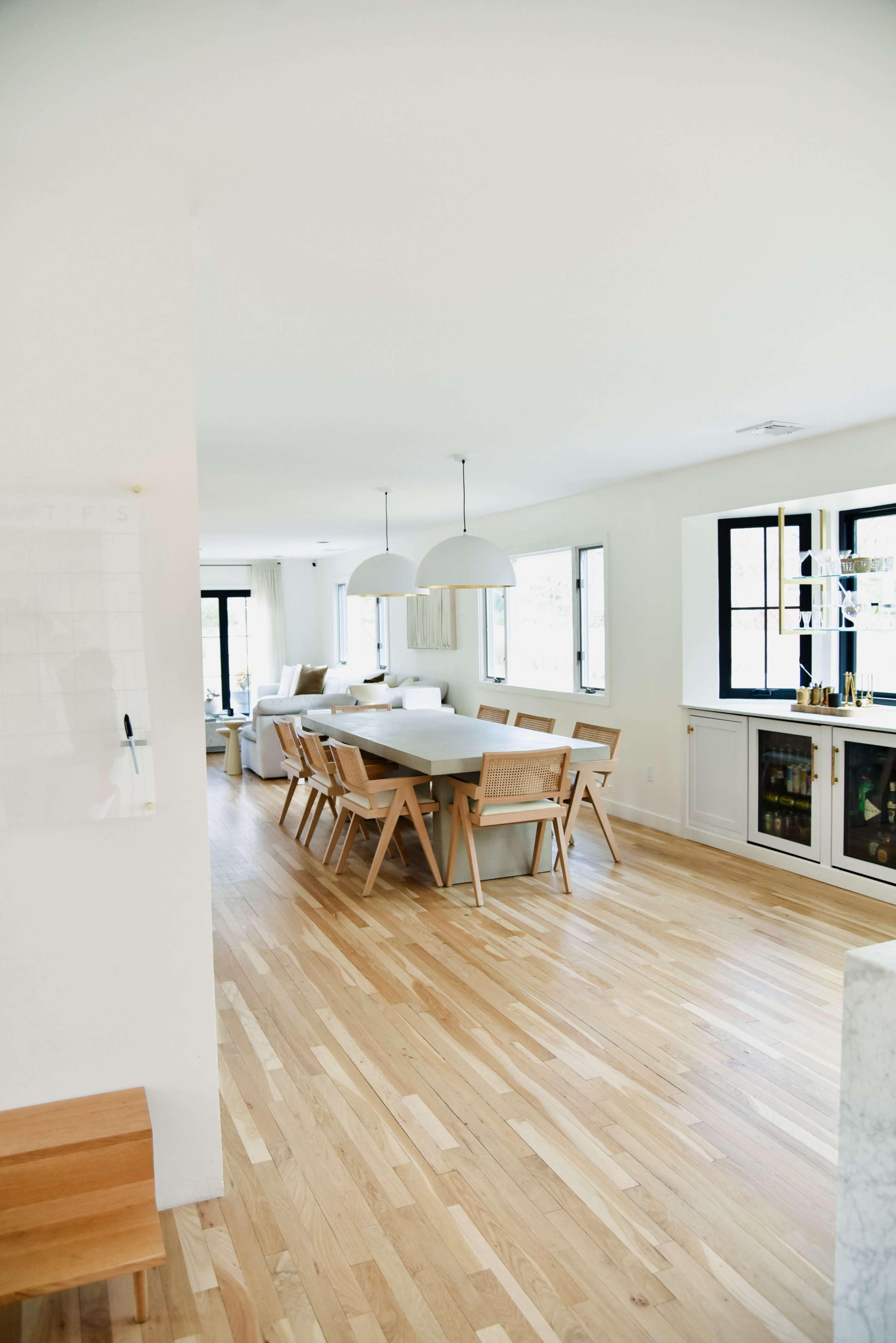 A modern, open-concept dining area features a long wooden table surrounded by light-colored chairs, with large windows allowing natural light to fill the space.