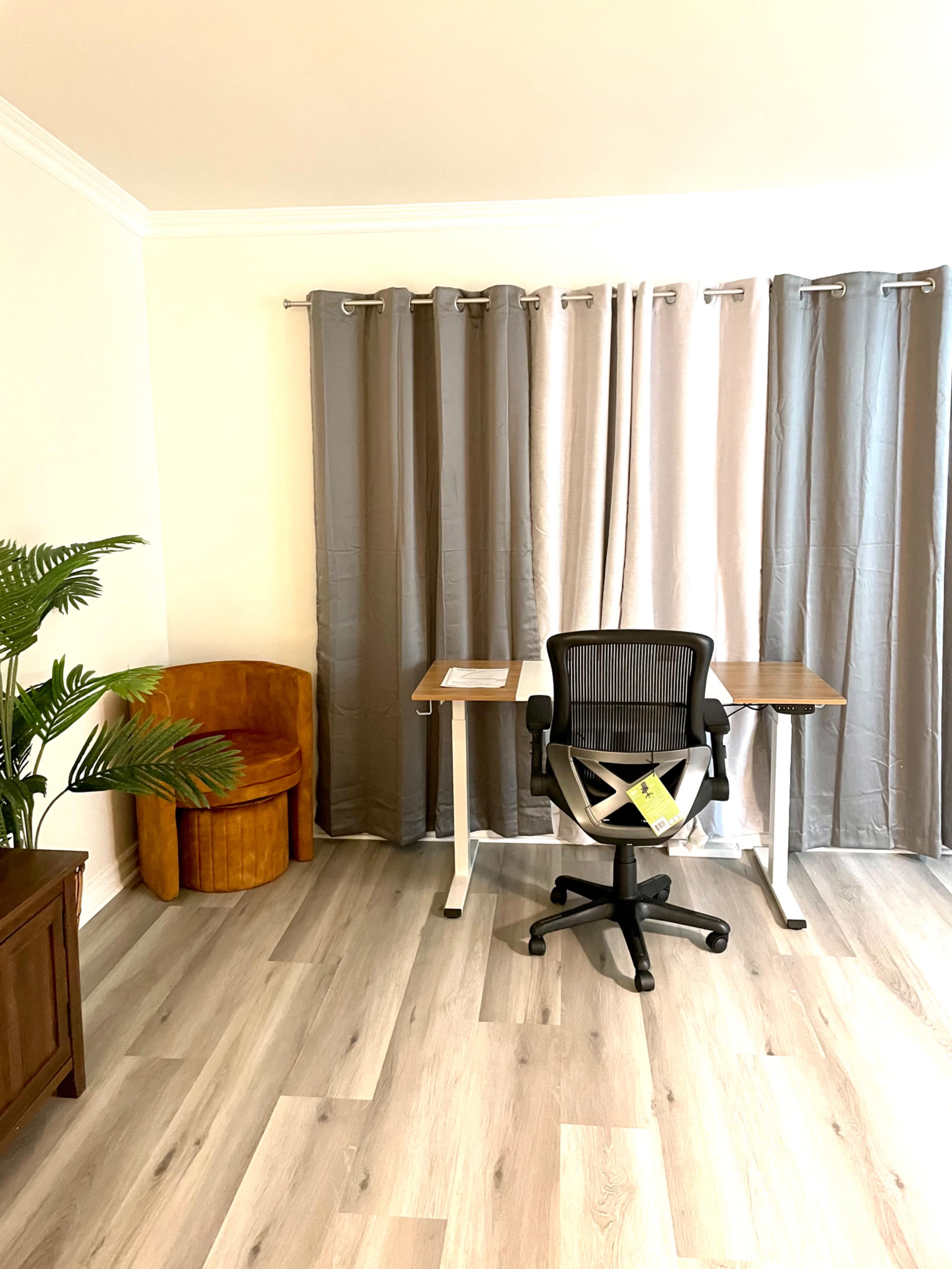 A modern home office features a desk with a chair, a wooden accent chair, and curtains in neutral tones.