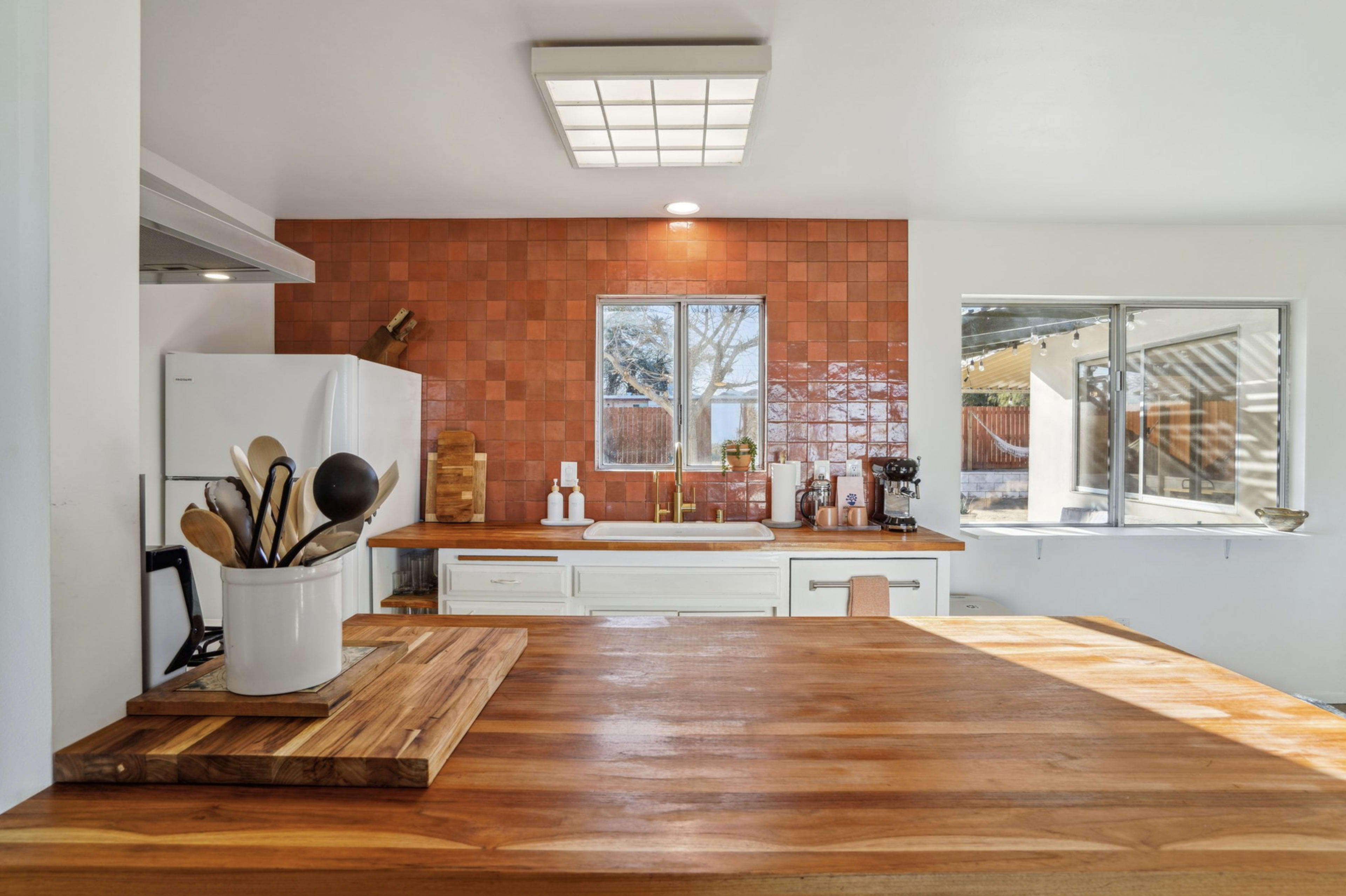 A modern kitchen features a large wooden island, a white refrigerator, and a wall of red tile behind a window.