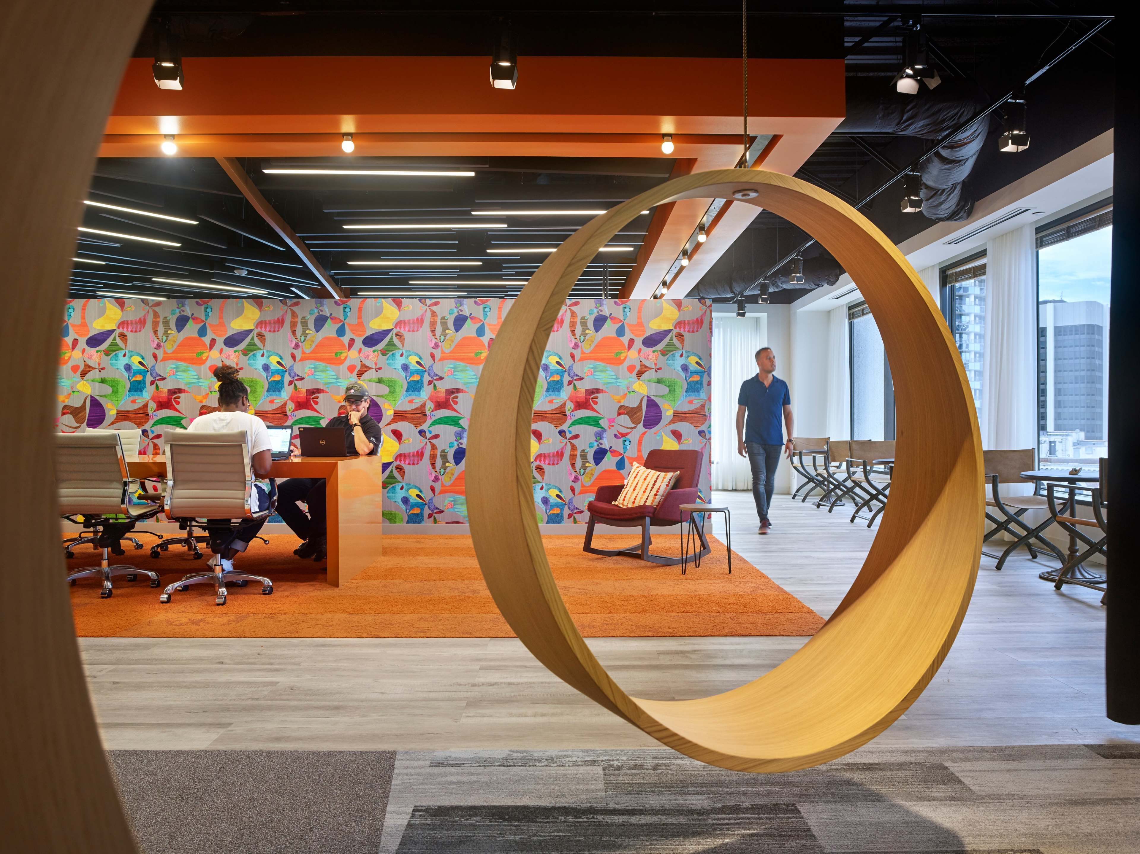 A modern office space with colorful wall art, a circular wooden element, and people engaged in conversation around a desk.