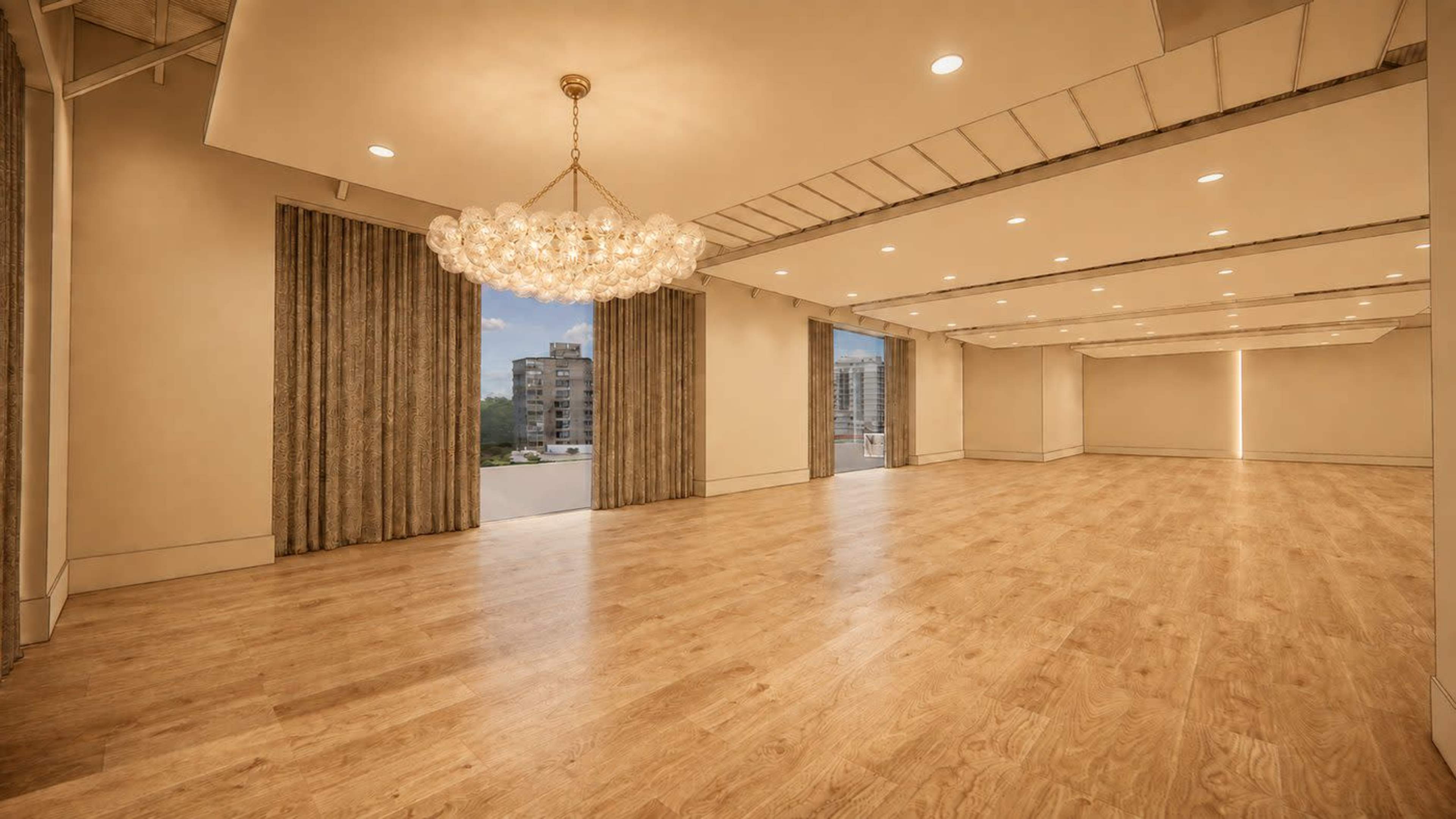 The image shows a spacious interior room with wooden flooring, a chandelier, and large windows that offer a view of an urban landscape.