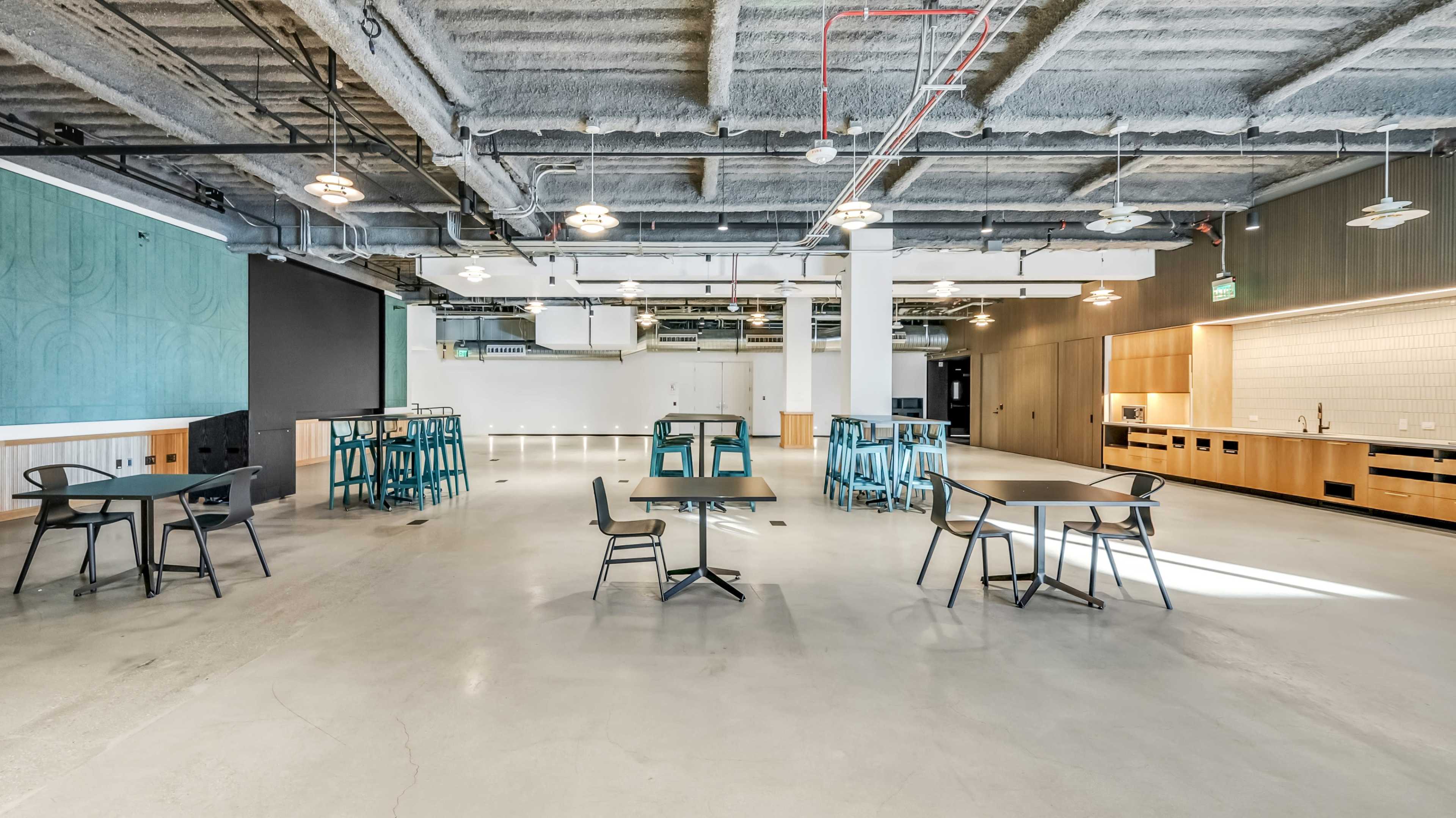 A spacious, modern dining area with several tables and chairs arranged on a concrete floor, large windows allowing natural light, and a kitchen area in the background.