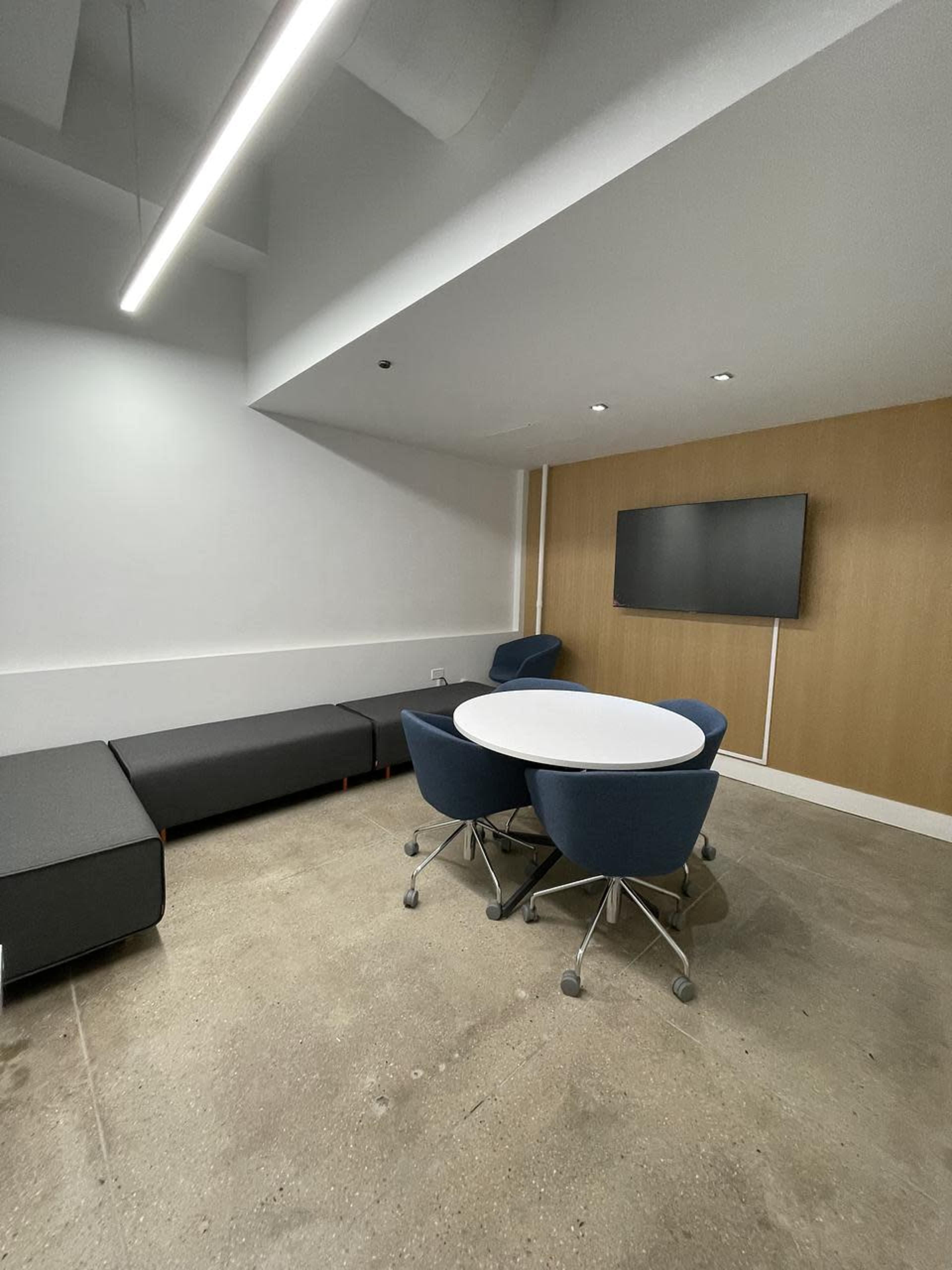 The image shows a modern conference room featuring a round table with four blue chairs, a gray couch along the wall, and a flat-screen TV mounted on a wooden panel.