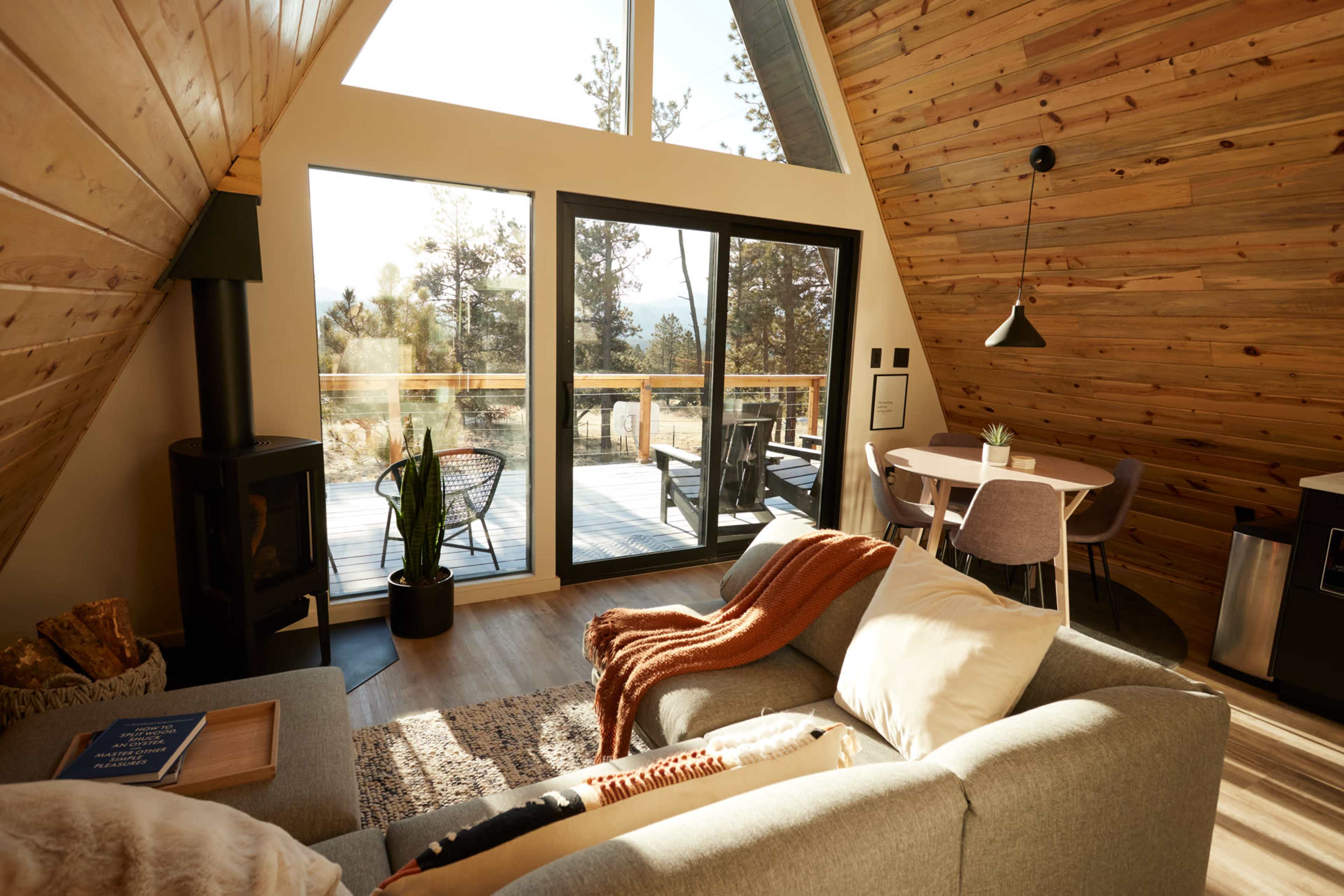 The interior of a modern A-frame cabin features a cozy seating area, large windows that overlook a wooden deck, and a dining space with minimalistic furniture.