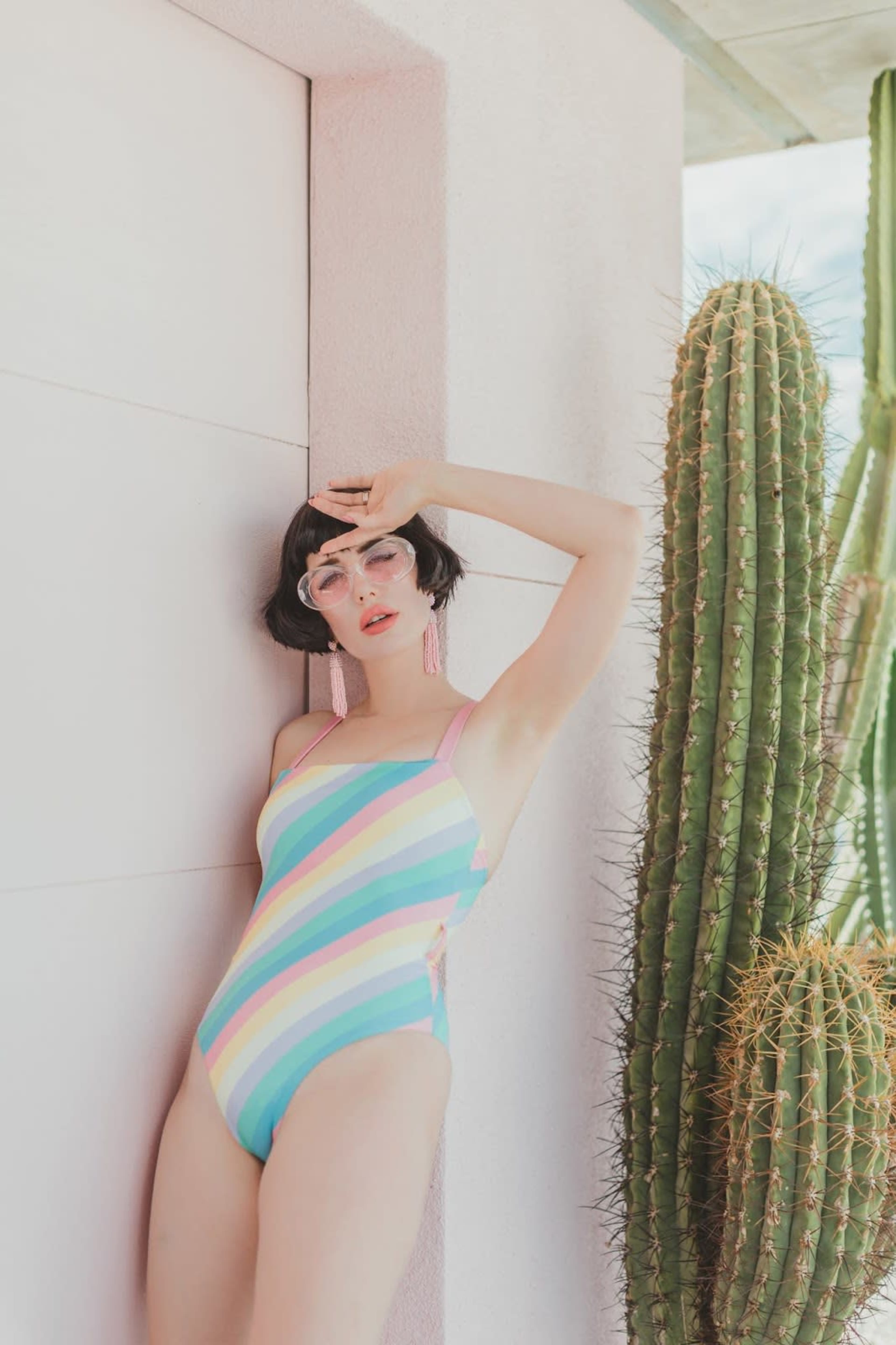 A woman in a striped swimsuit stands beside a tall cactus against a white wall.