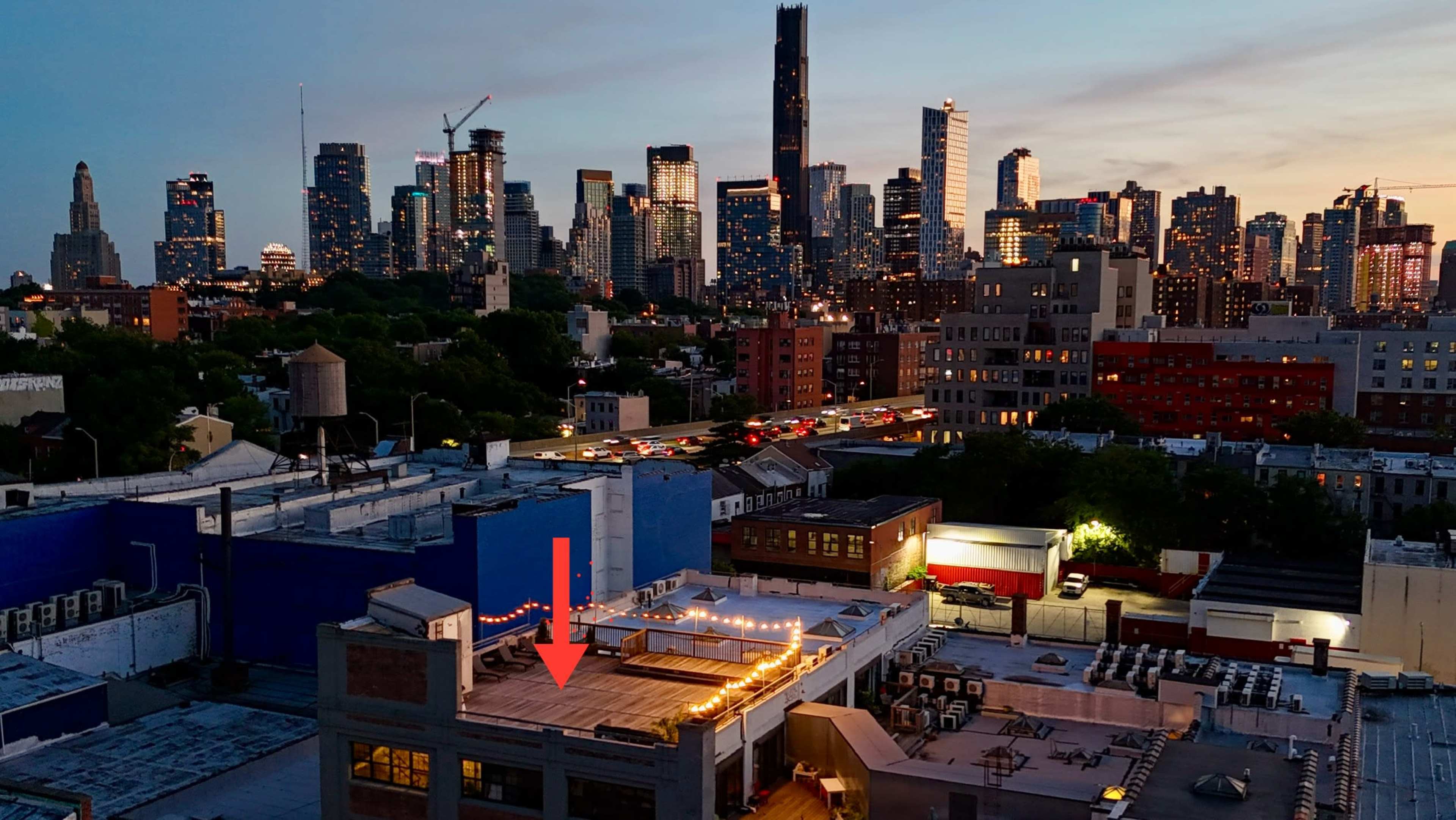 A city skyline at dusk with skyscrapers illuminated against a fading sky, and a rooftop with string lights in the foreground.