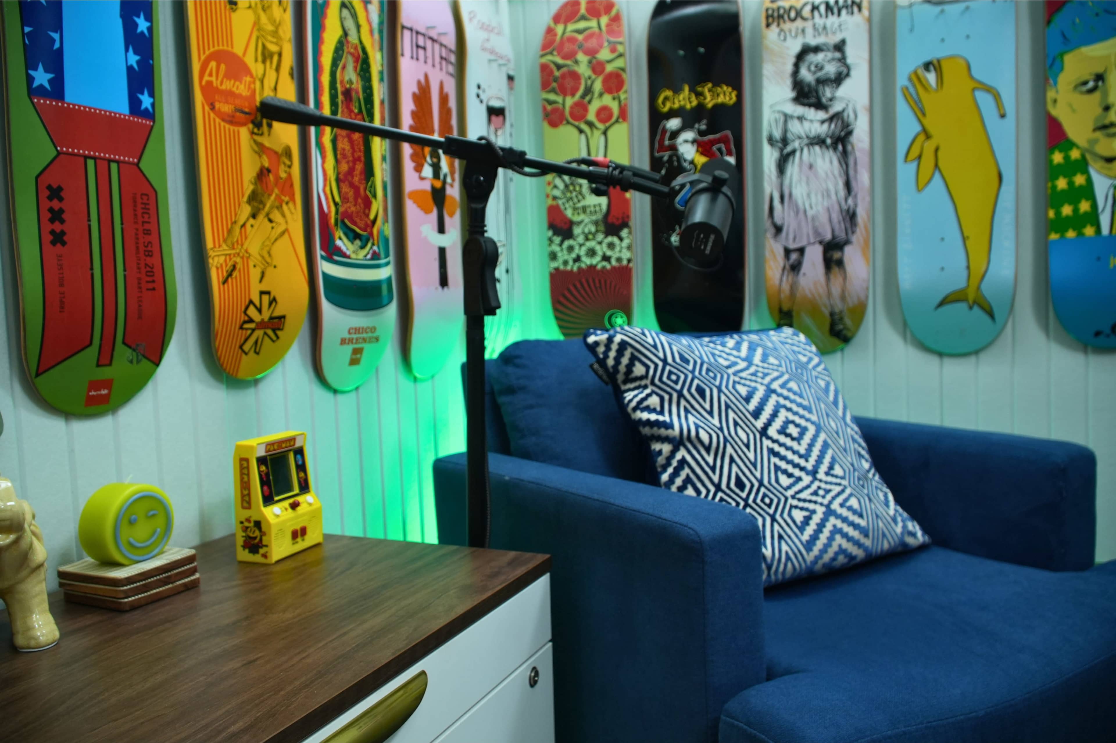 The image features a blue armchair beside a wooden table with a vintage arcade game, against a backdrop of colorful skateboards mounted on the wall.