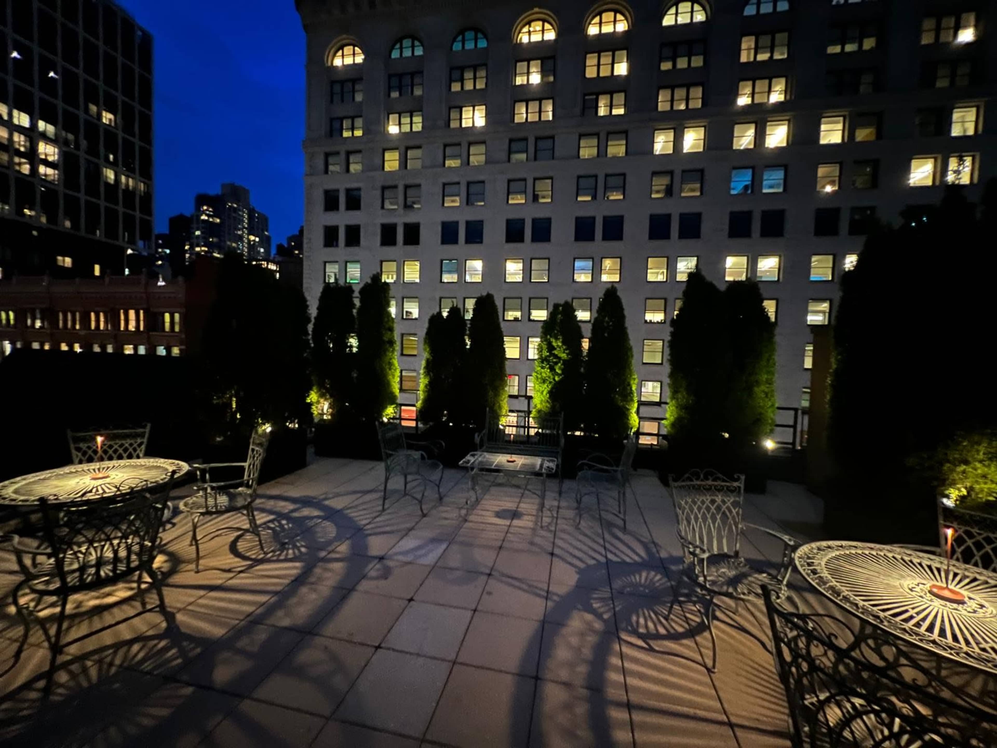 The image shows a rooftop terrace featuring several metal tables and chairs, illuminated by surrounding city lights at night.
