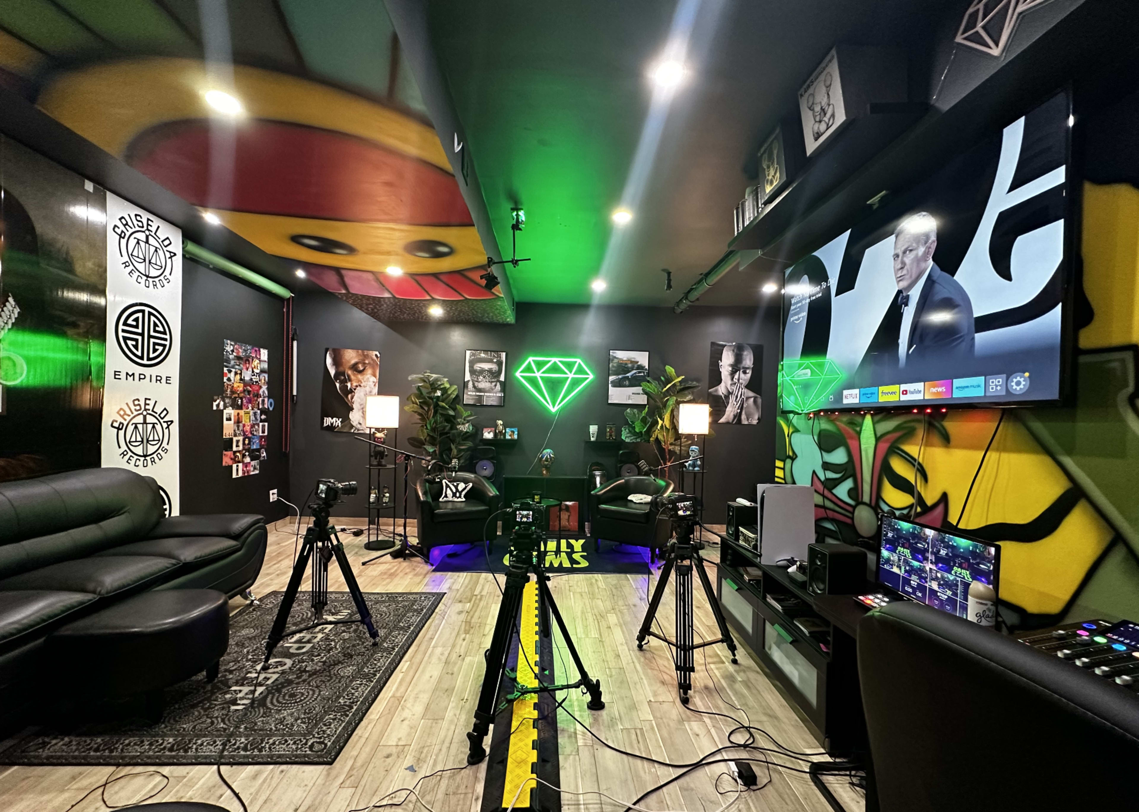 A modern multimedia studio setup with green lighting, cameras, and a large screen displaying a presentation, surrounded by vibrant wall art and photographs.