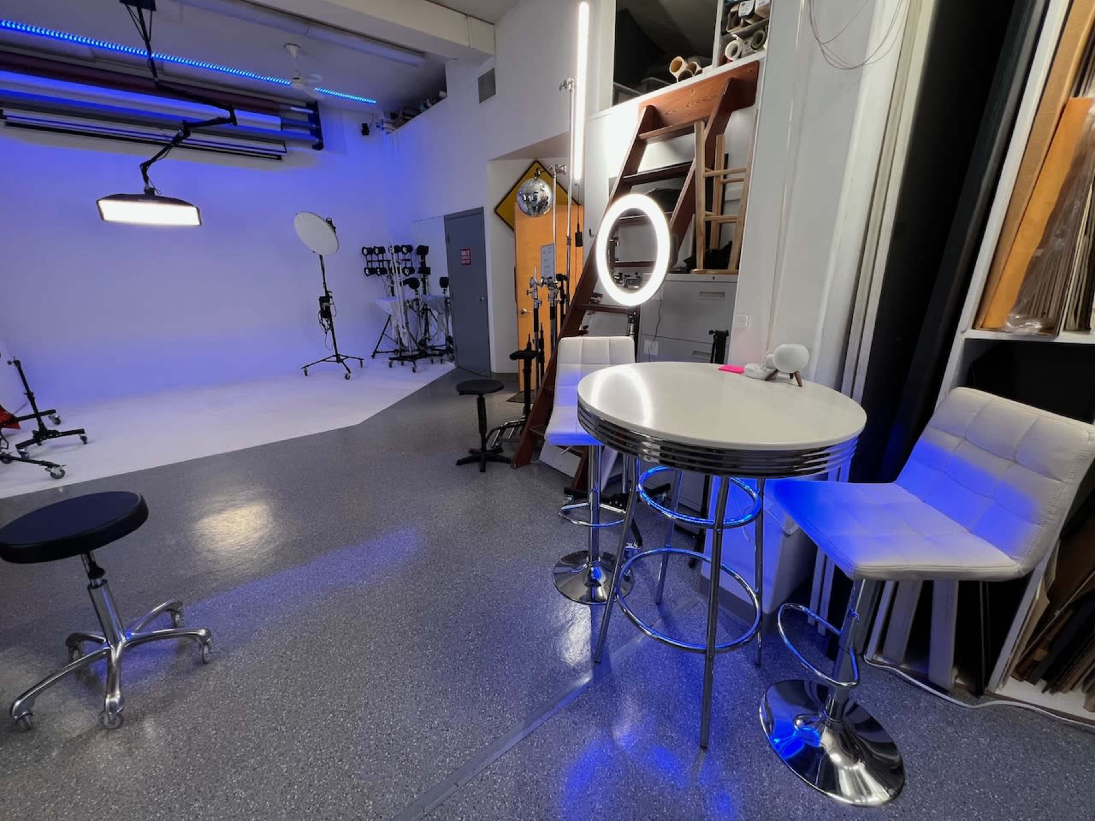 A photography studio with various lighting equipment, a round table with bar stools, and a mirror ring light, all set against a grey floor.