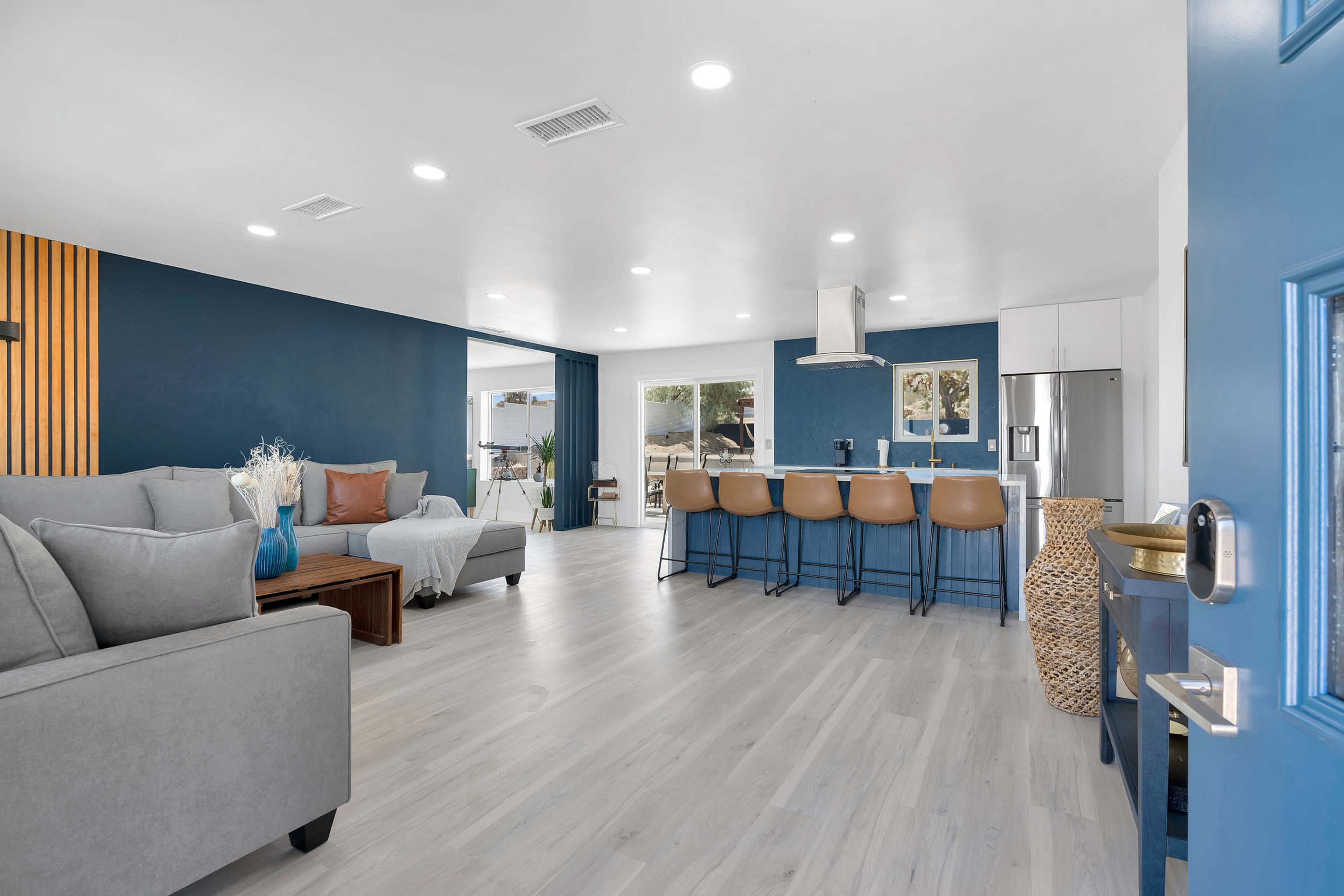 A modern living space featuring a gray sofa, a wooden coffee table, a kitchen area with bar stools, and blue accent walls.