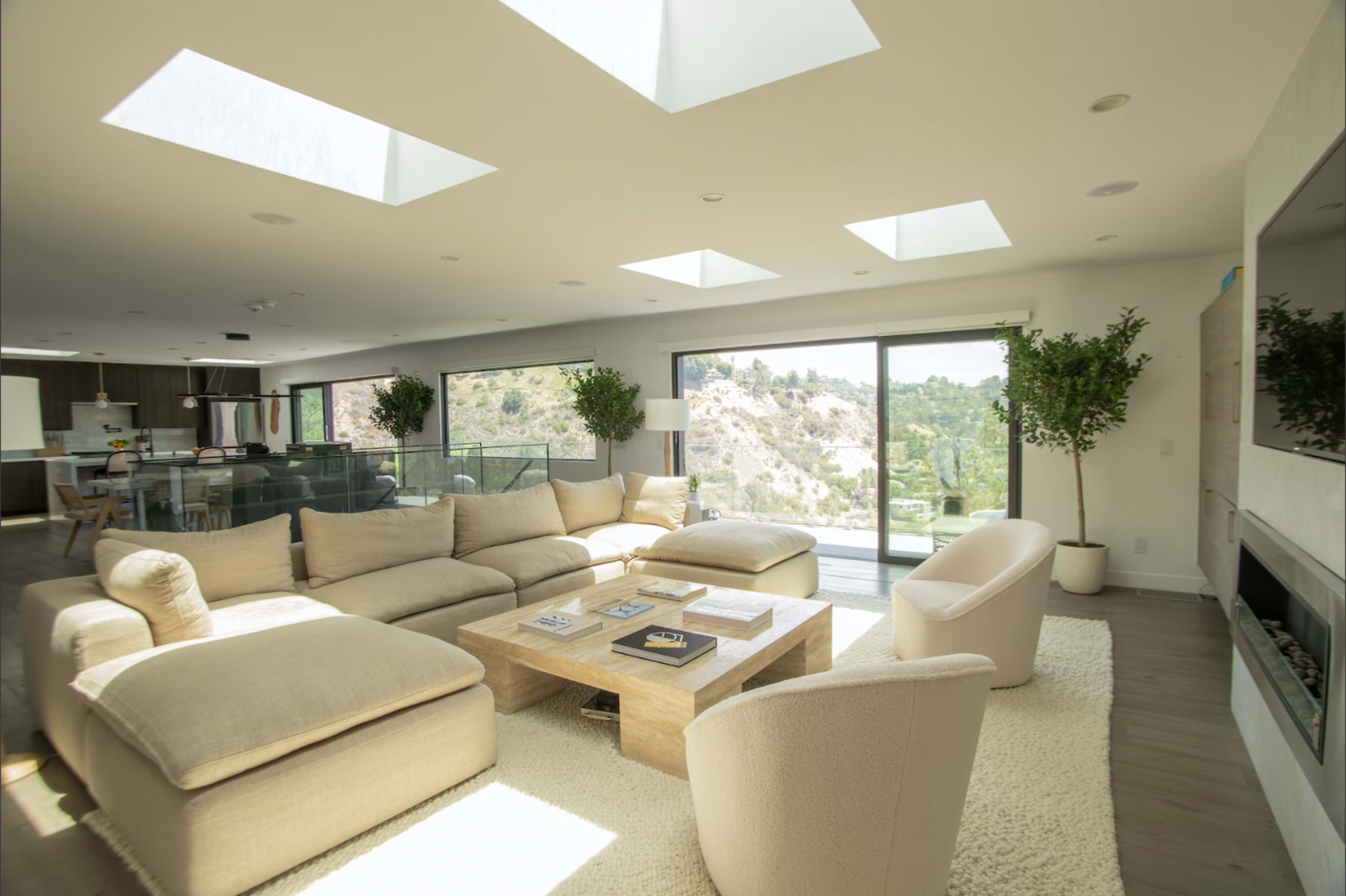 A modern living room features a large, beige sectional sofa, a wooden coffee table, and floor-to-ceiling windows with views of a hillside.