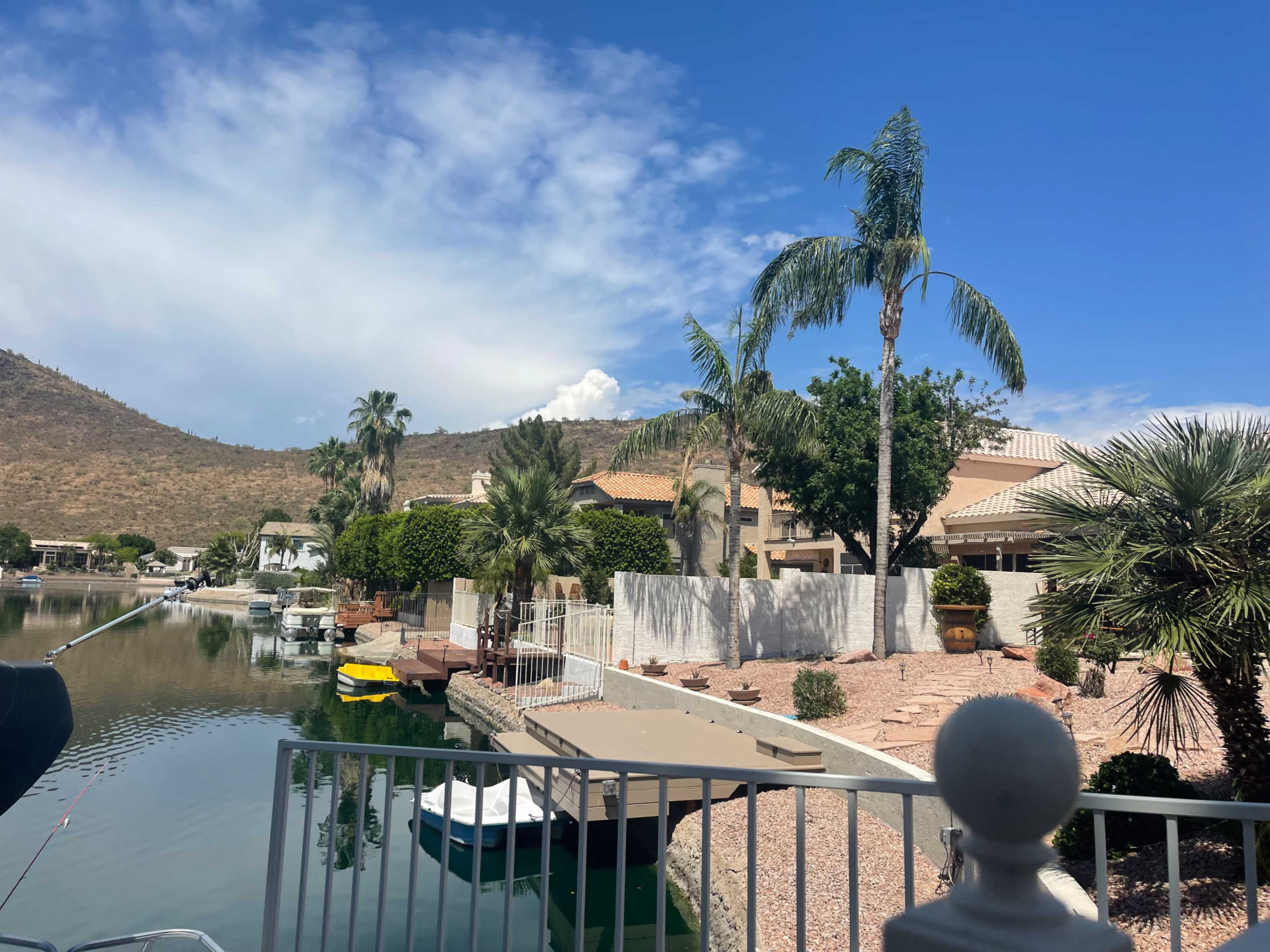 The image shows a waterfront scene with palm trees, residential buildings, and boats docked along a canal against a backdrop of hills and blue sky.
