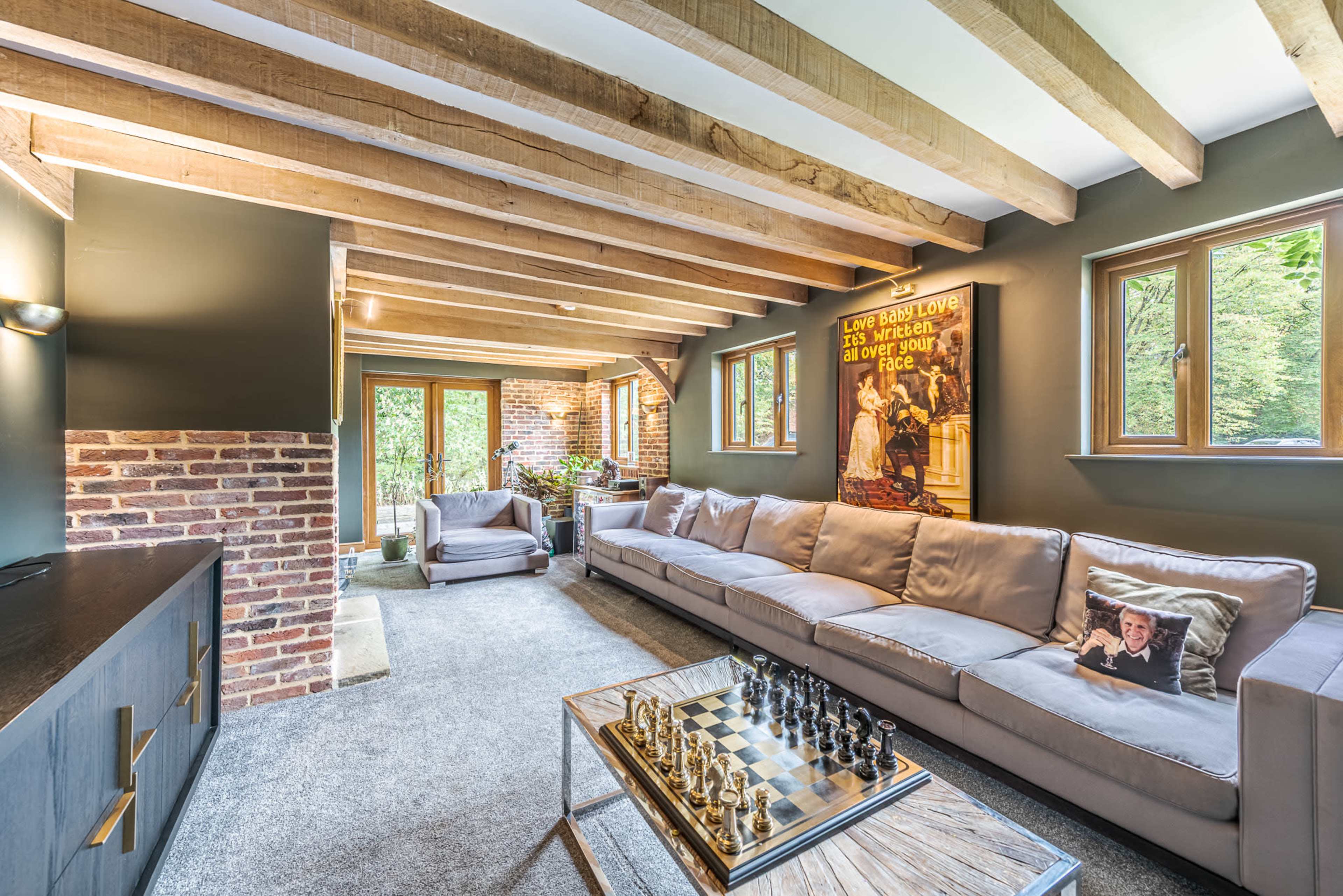 The image shows a cozy living room with a large sectional sofa, a chess set on a glass coffee table, exposed wooden beams, and windows letting in natural light.