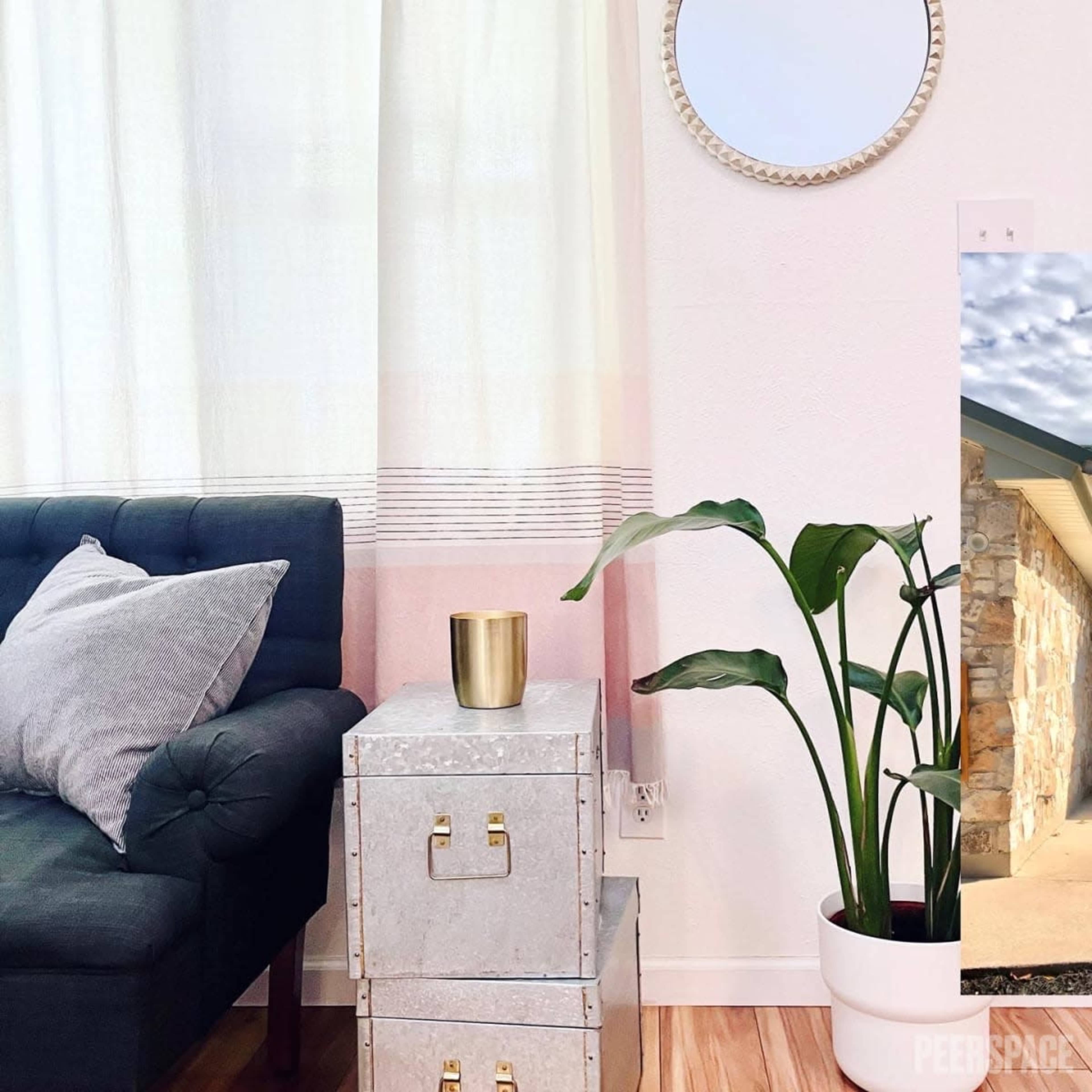 A cozy living space features a dark grey sofa, a decorative mirror, a potted plant, and stacked metallic storage boxes by a sheer curtain.
