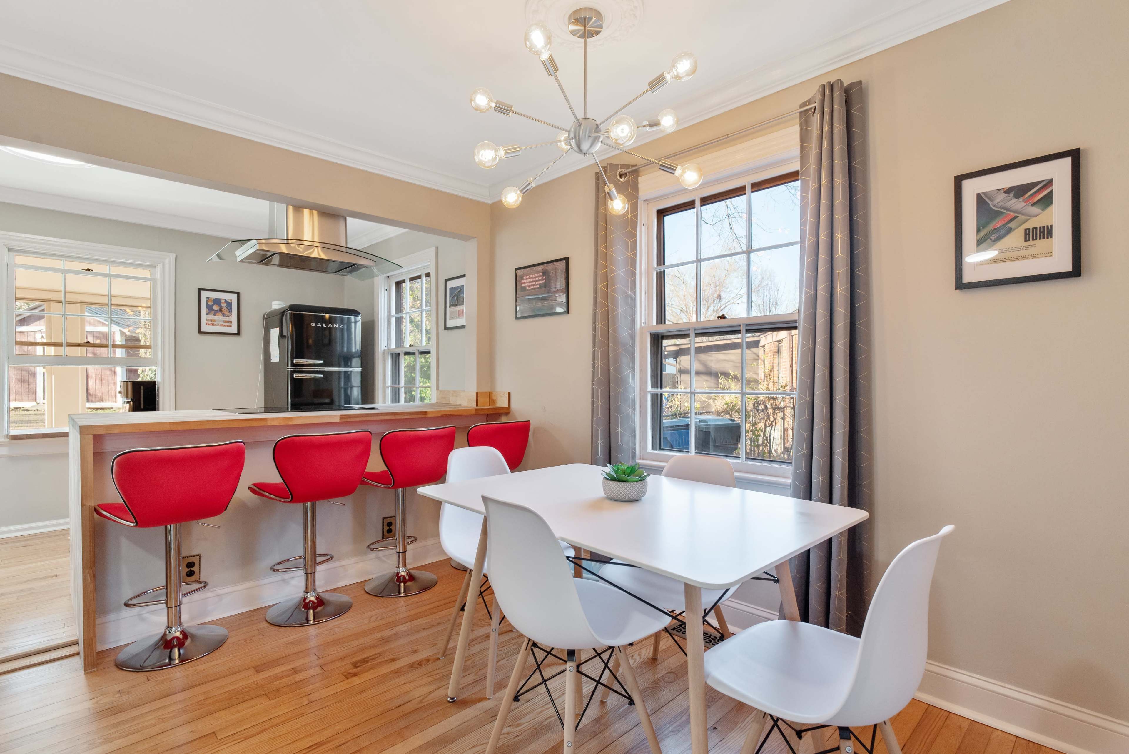 A modern dining area with a white table and chairs beside a kitchen bar with red stools, illuminated by a contemporary light fixture.