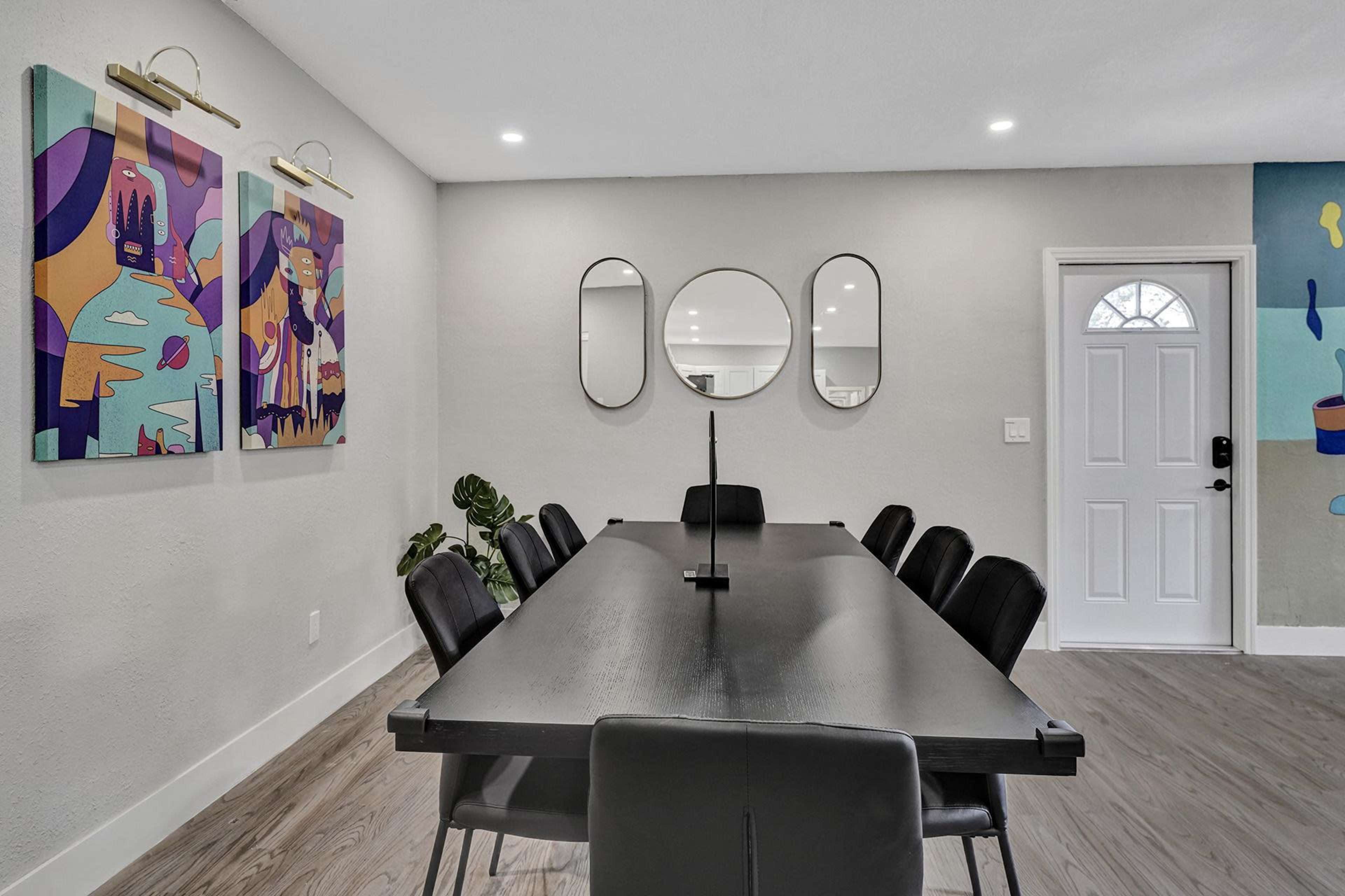 A modern dining area featuring a long black table with eight chairs, illuminated by wall art and two mirrors, adjacent to a white door.