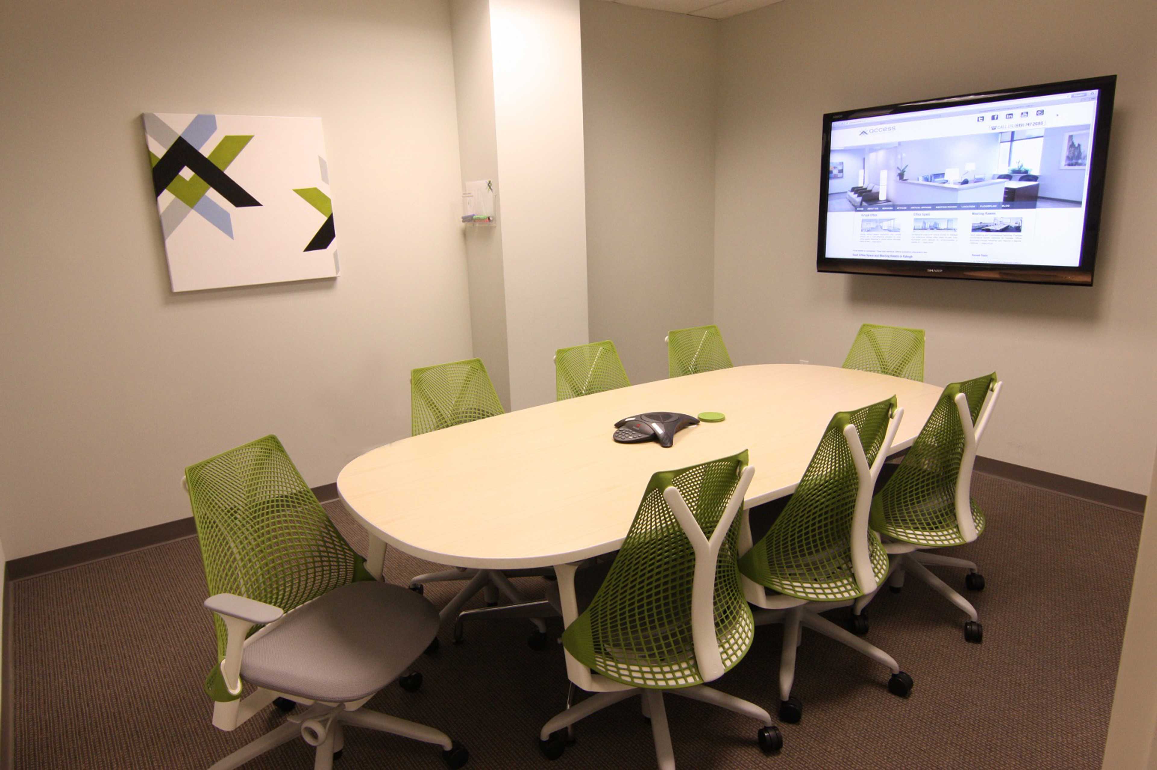A modern conference room features a large oval table surrounded by green mesh chairs and a wall-mounted screen displaying a webpage.