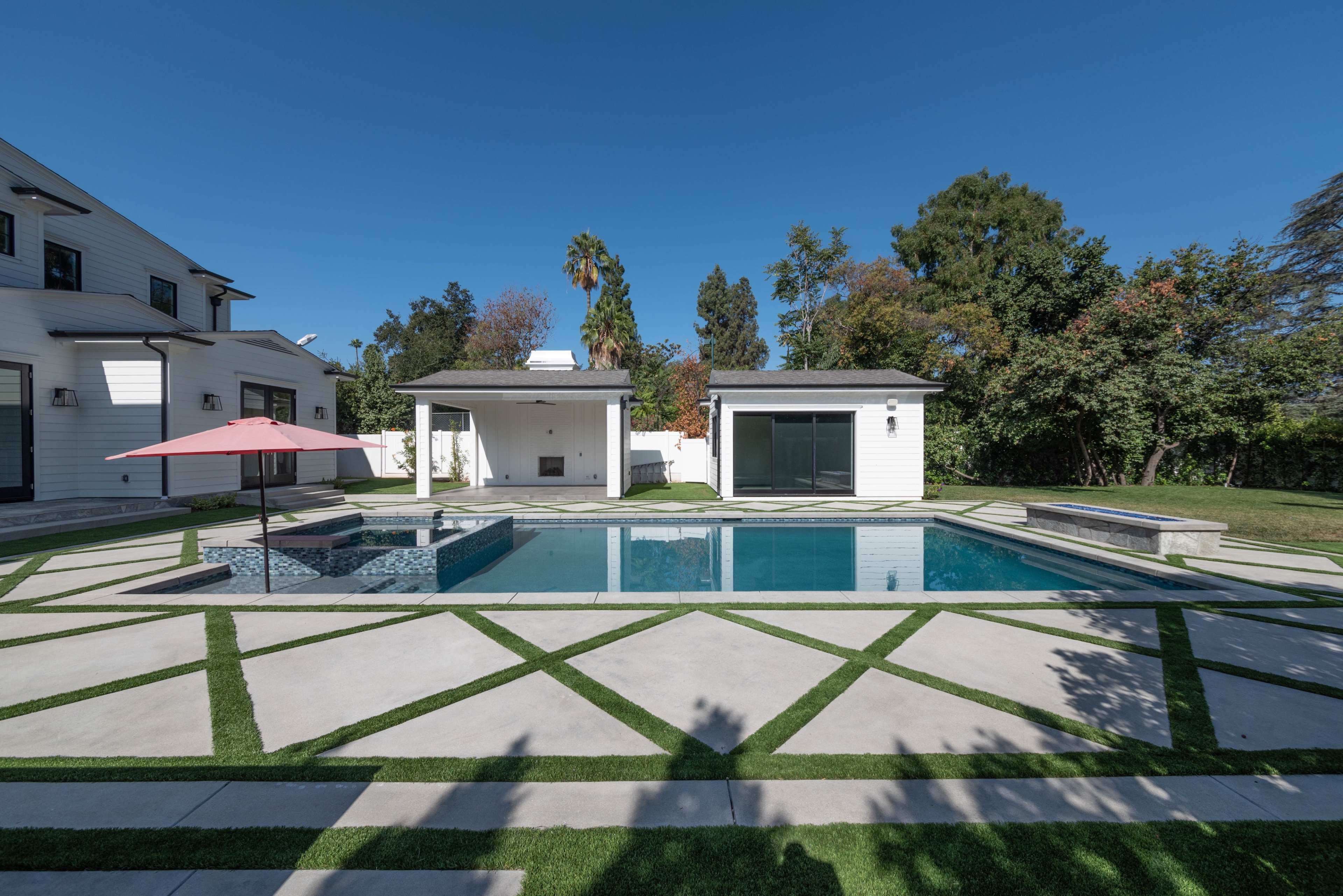 The image shows a modern backyard featuring a swimming pool surrounded by geometric stone pathways and landscaped greenery, with a shaded seating area and poolside structures.