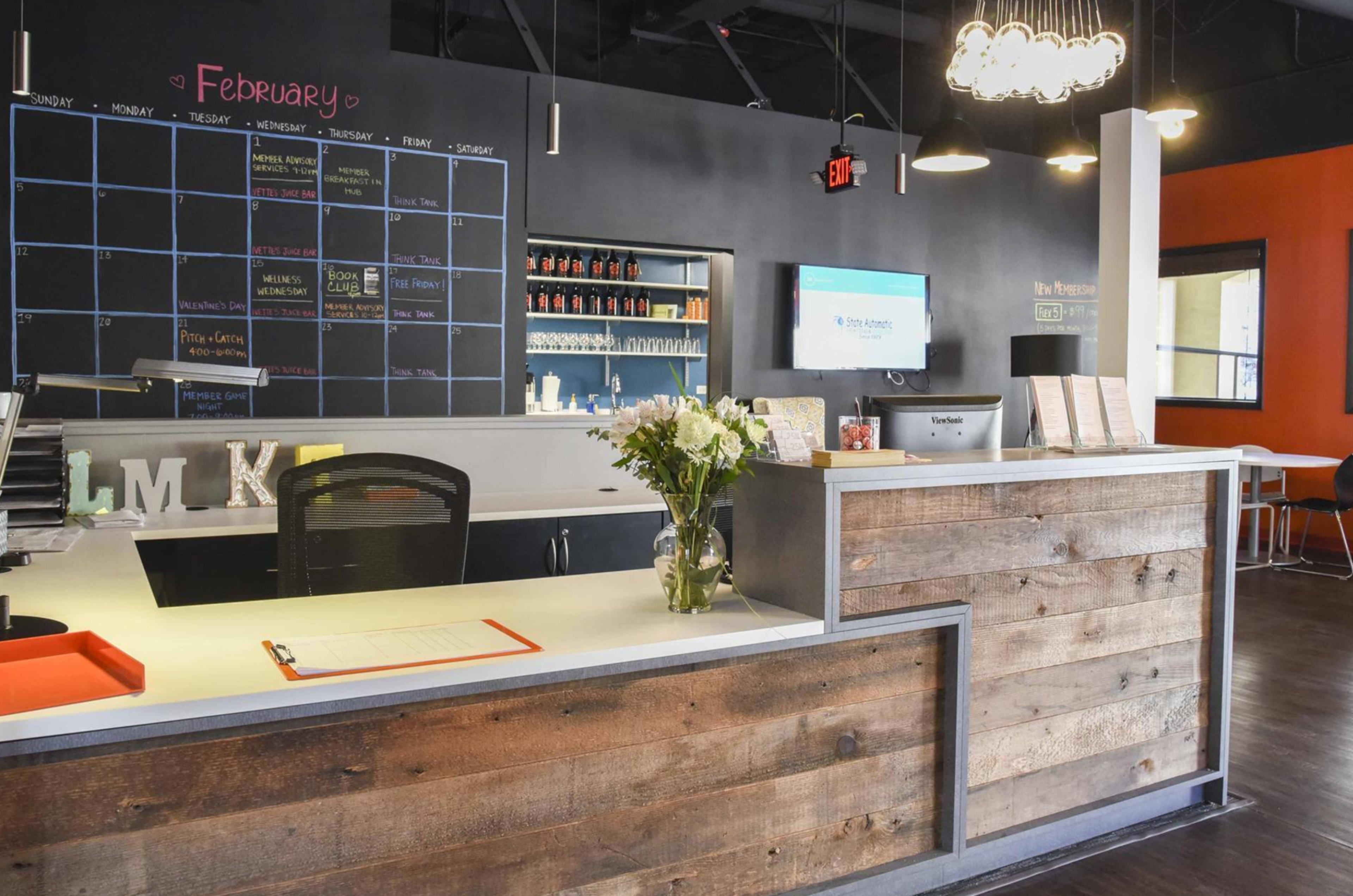A modern reception area with a wooden front desk, a calendar on the wall, and a floral arrangement on the counter.