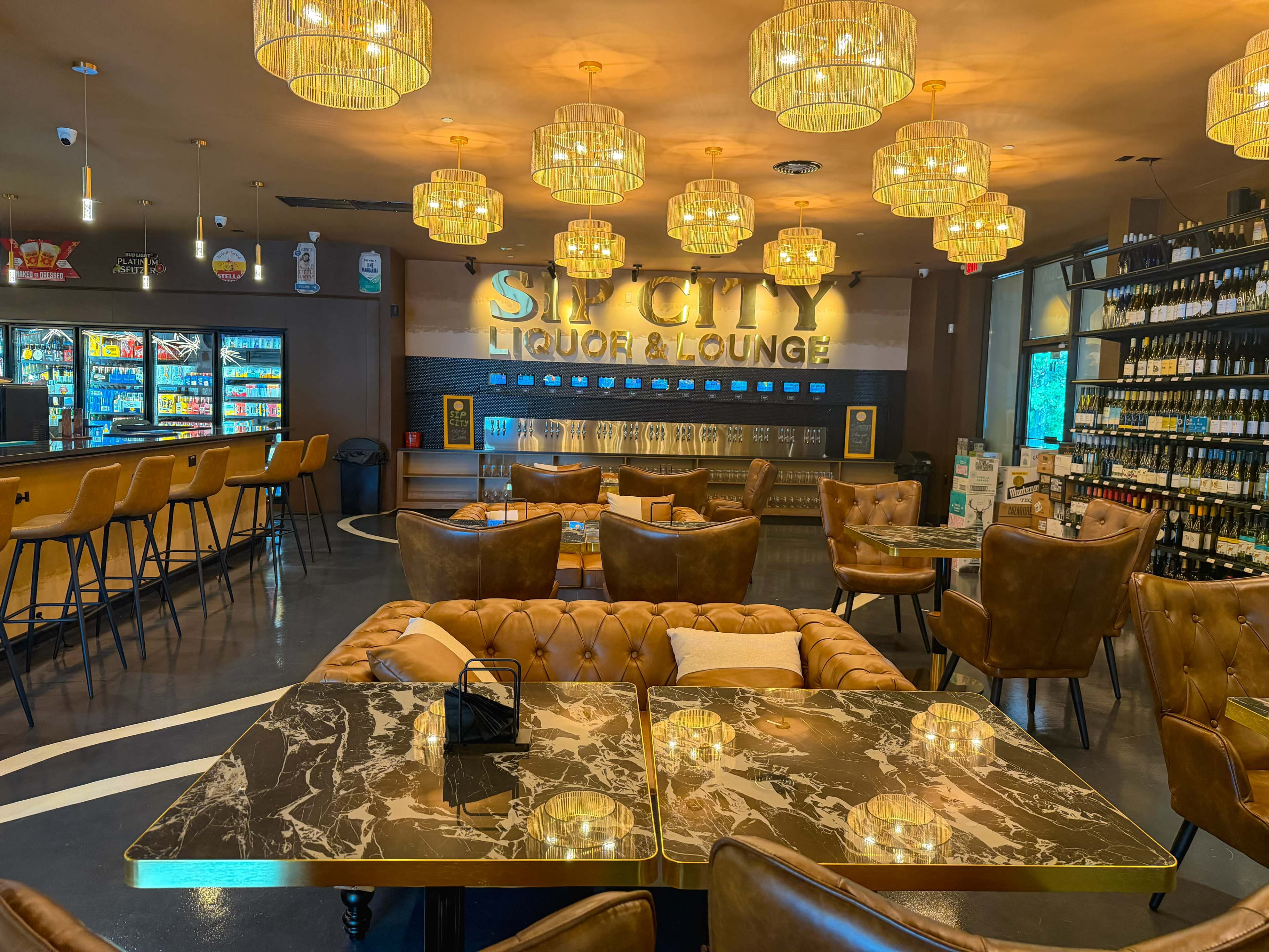 The image shows an upscale lounge with plush seating, a marble table, and a well-stocked bar featuring a variety of beverages.