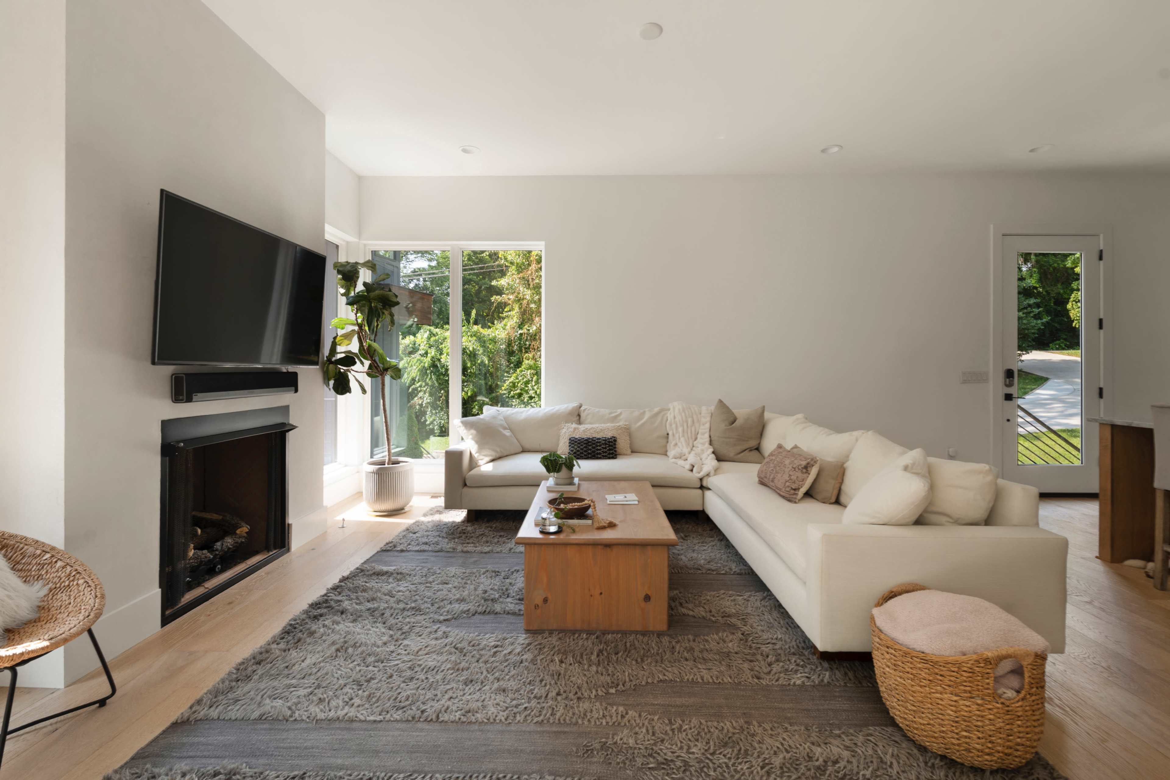 The image shows a bright, modern living room with a large white sectional sofa, a wooden coffee table, a TV mounted on the wall, and large windows allowing natural light.