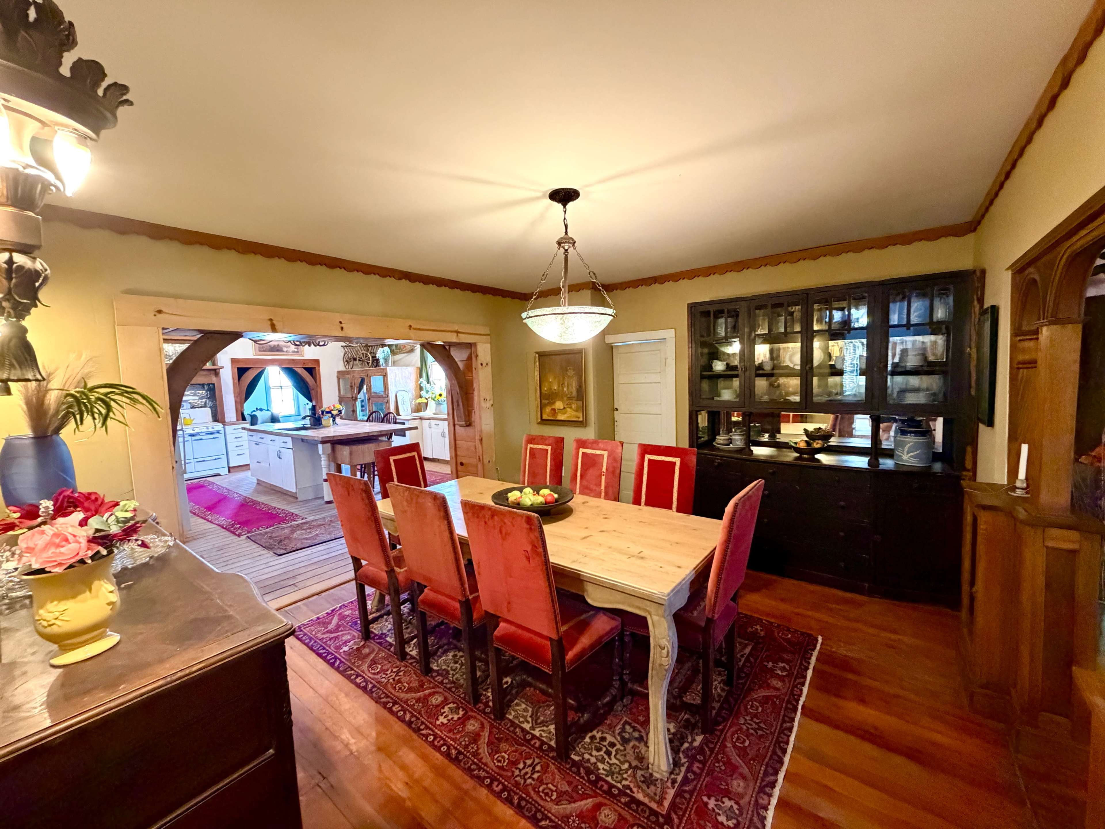 The image shows a dining room with a long wooden table surrounded by red chairs, a sideboard with glass doors, and an open entrance leading to a kitchen area.