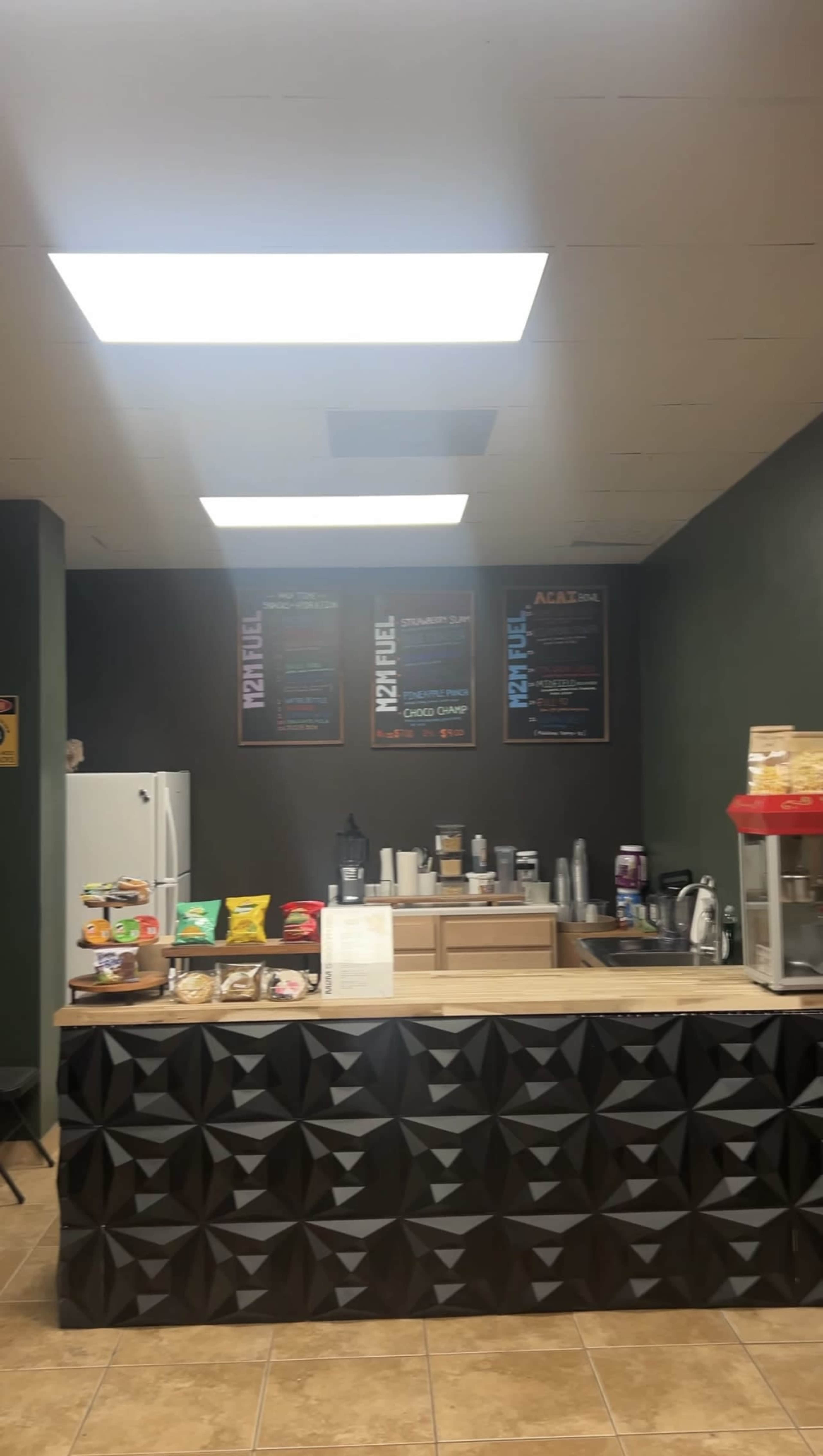 The image shows a small cafe with a counter displaying snacks and beverages, along with a menu on the wall and a refrigerator in the background.
