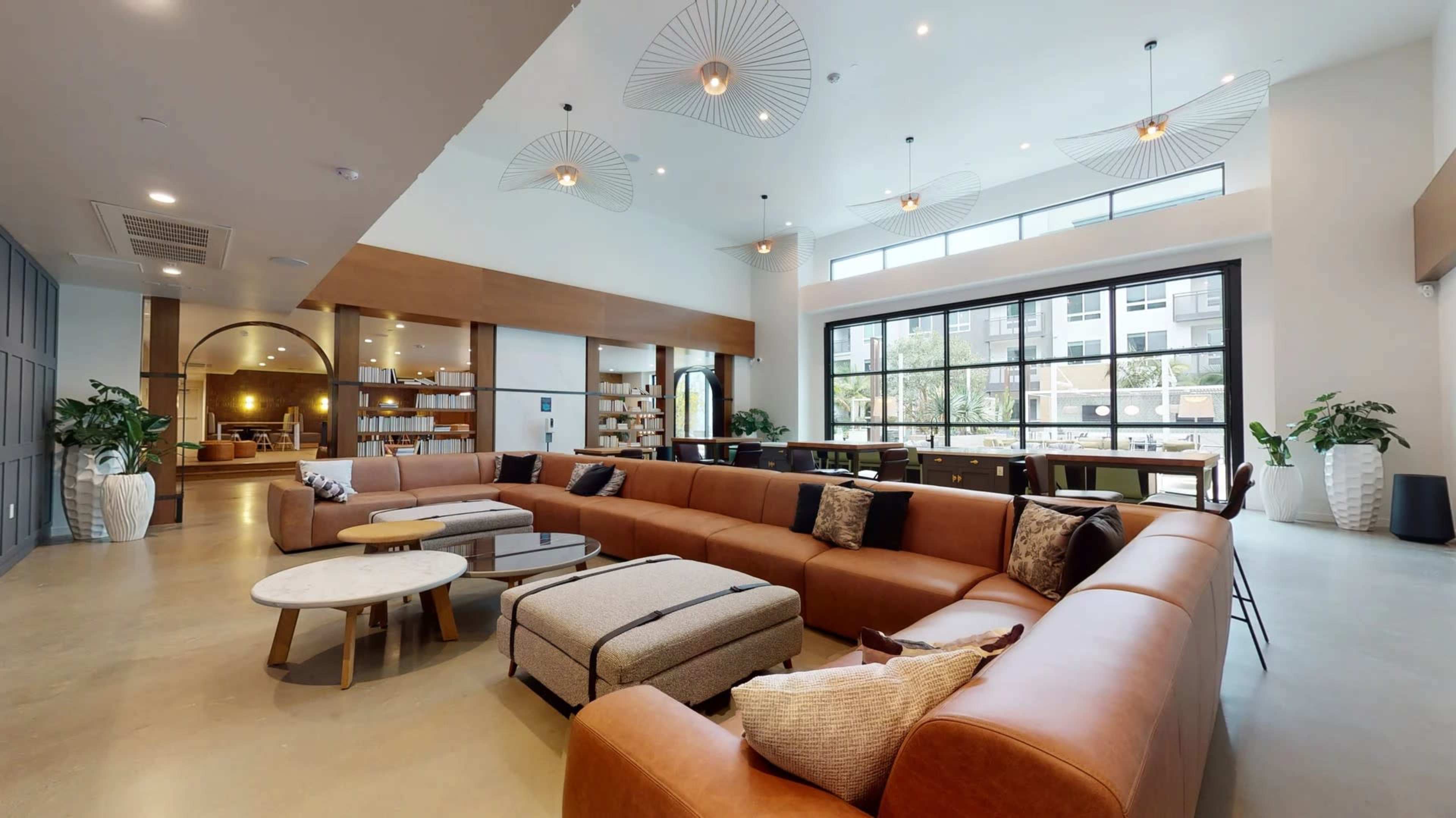 A spacious modern lounge area features a large brown sectional sofa, round coffee tables, and ample natural light from large windows.
