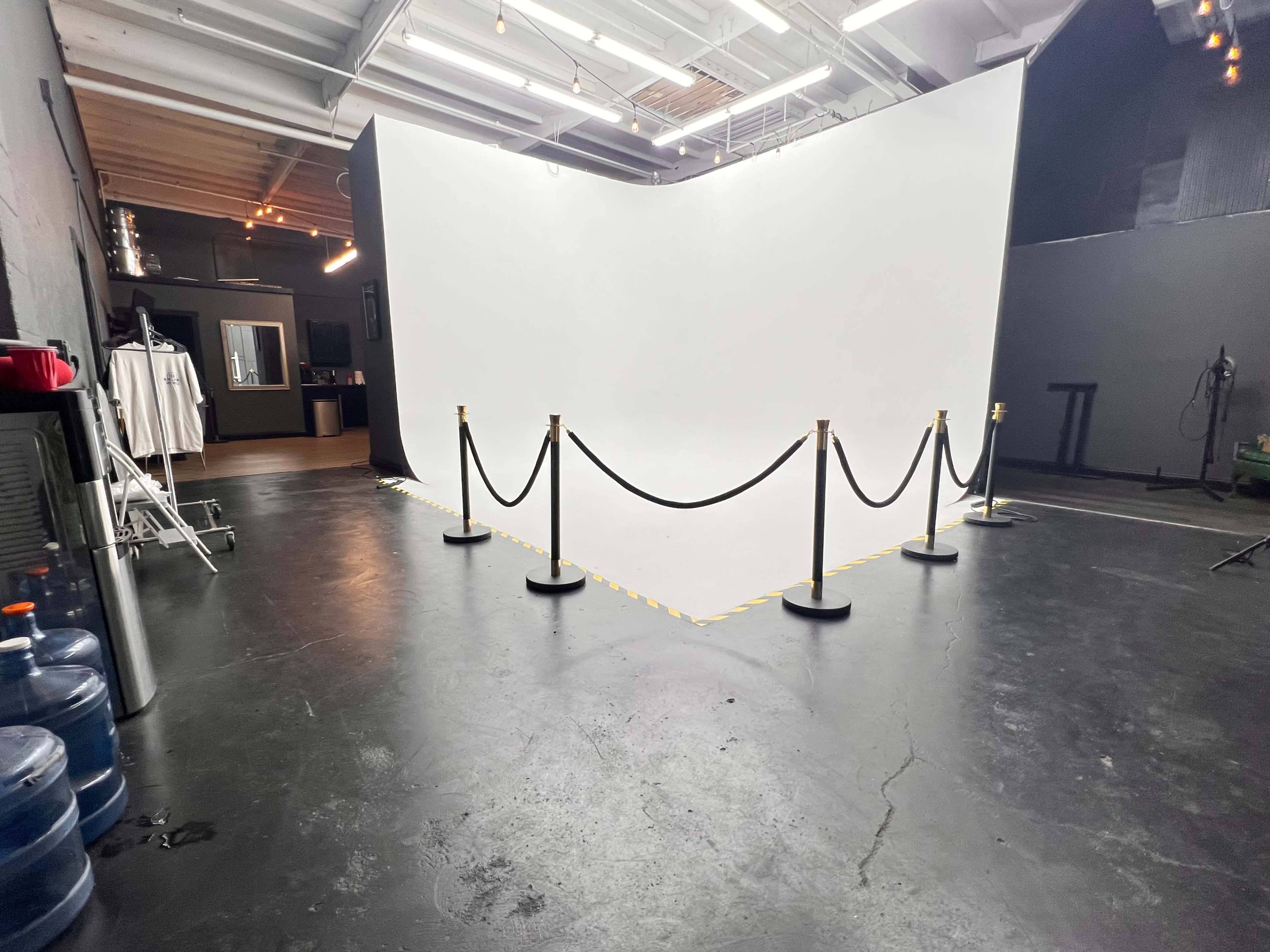 The image shows a photography studio with a large white backdrop, surrounded by a rope barrier and equipped with water dispensers in the corner.