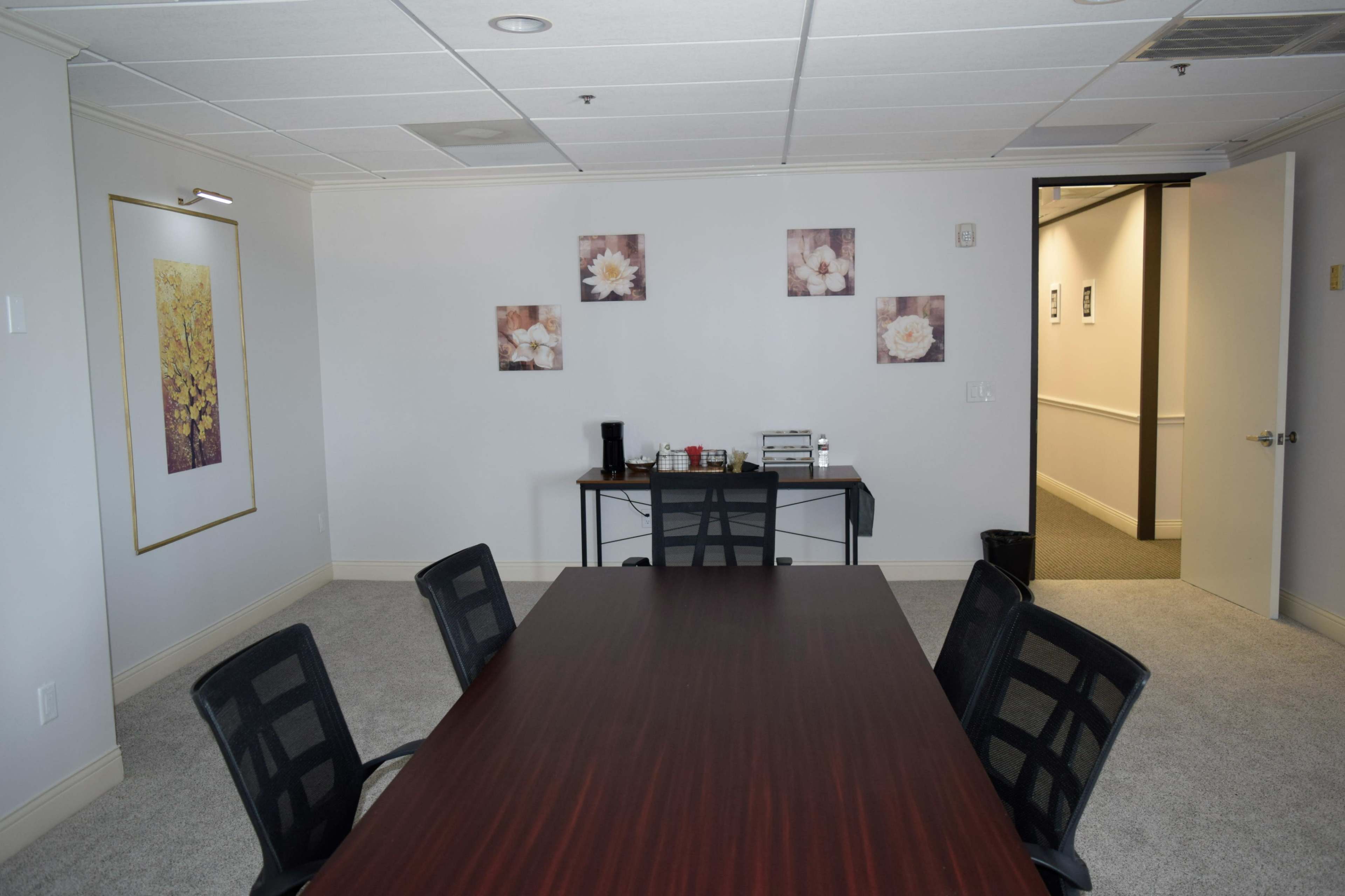 A conference room with a wooden table, black chairs, and floral artwork on the walls, leading to an open doorway.