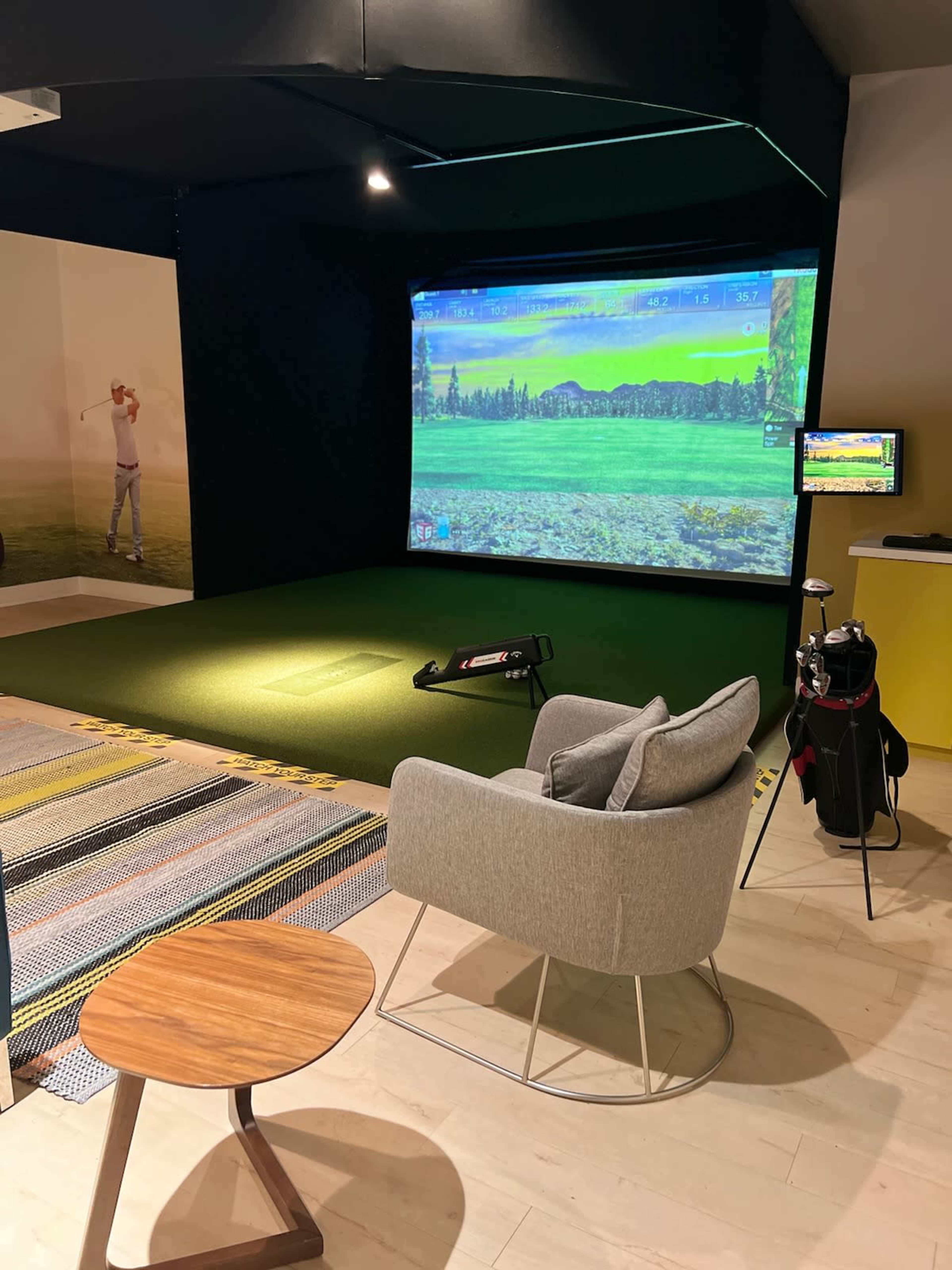 An indoor golf simulation room featuring a large projection screen displaying a golf course scene, a turf area for practice, a golf bag, a chair, and a small table.