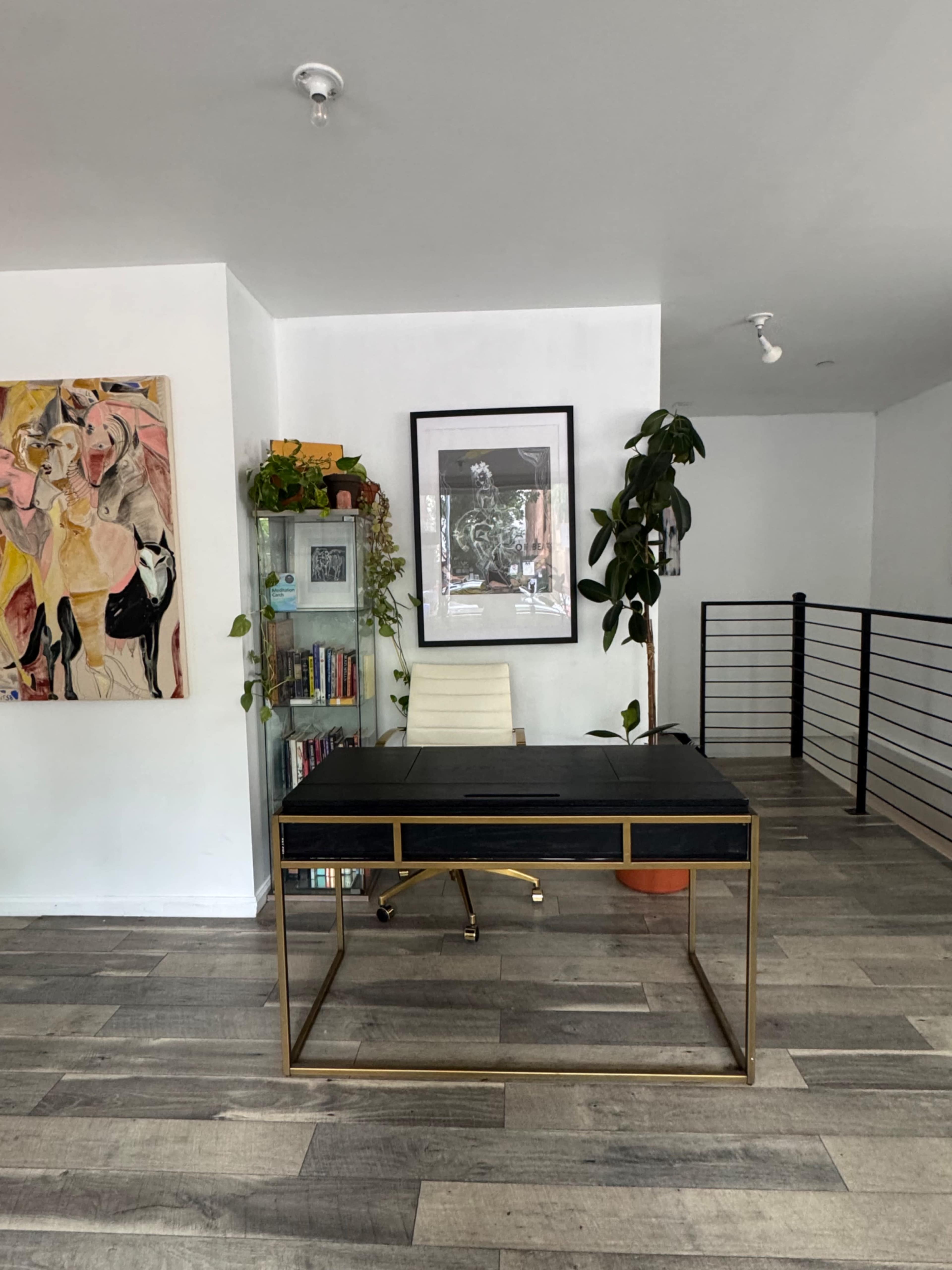 The image shows a modern home office with a black desk, a white chair, a bookshelf, a plant, and artwork on the wall.