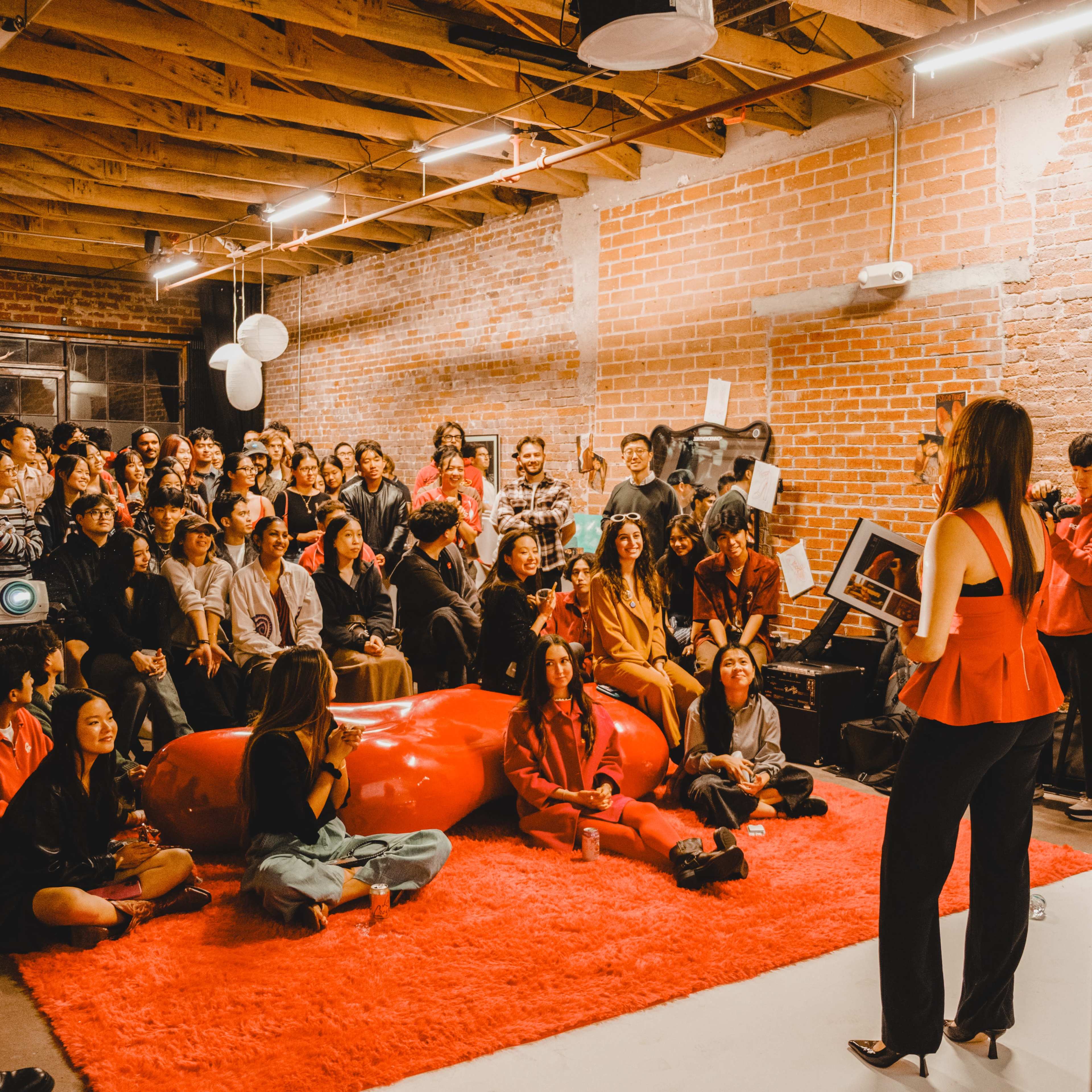 A crowd of people sits on a red carpet in a brick-walled room, attentively watching a speaker holding a book.