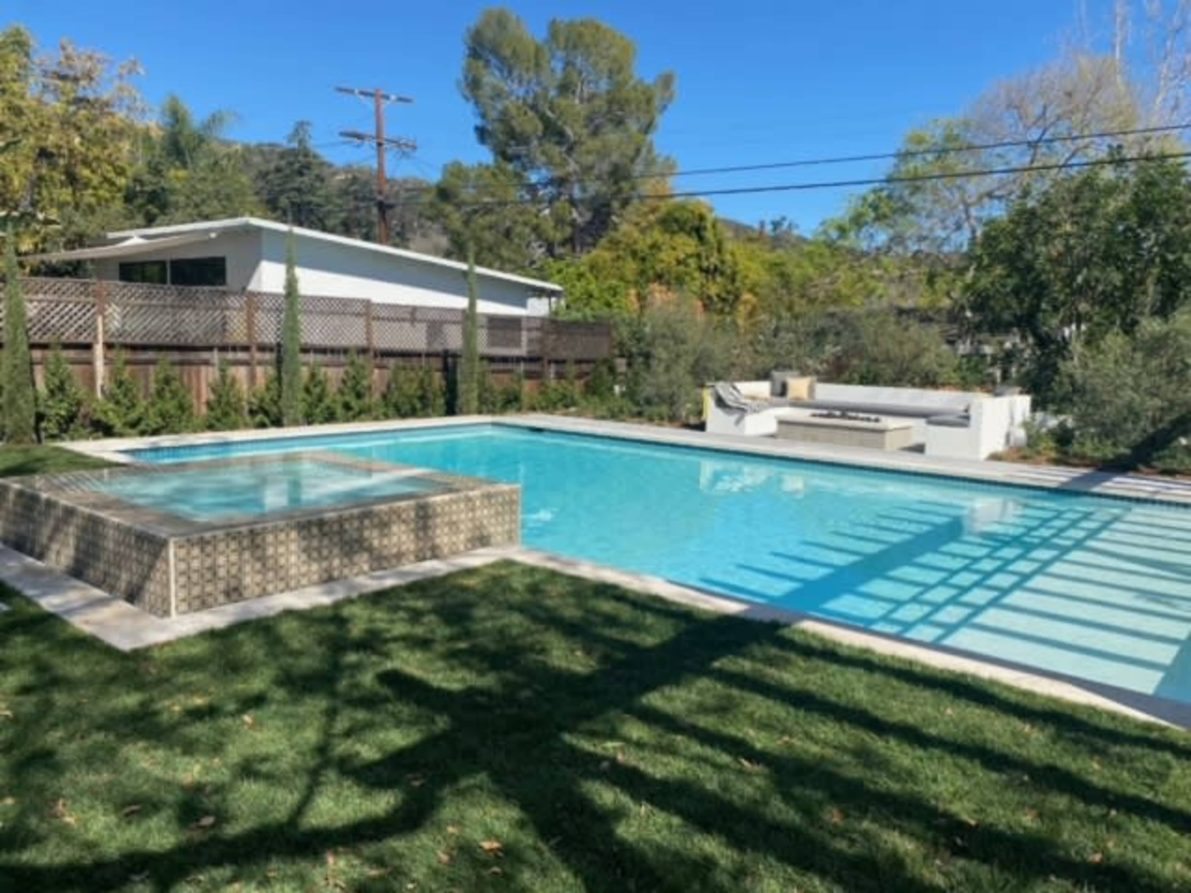 A rectangular swimming pool surrounded by a grassy area and modern landscaping, with a raised spa structure at one end and outdoor seating nearby.