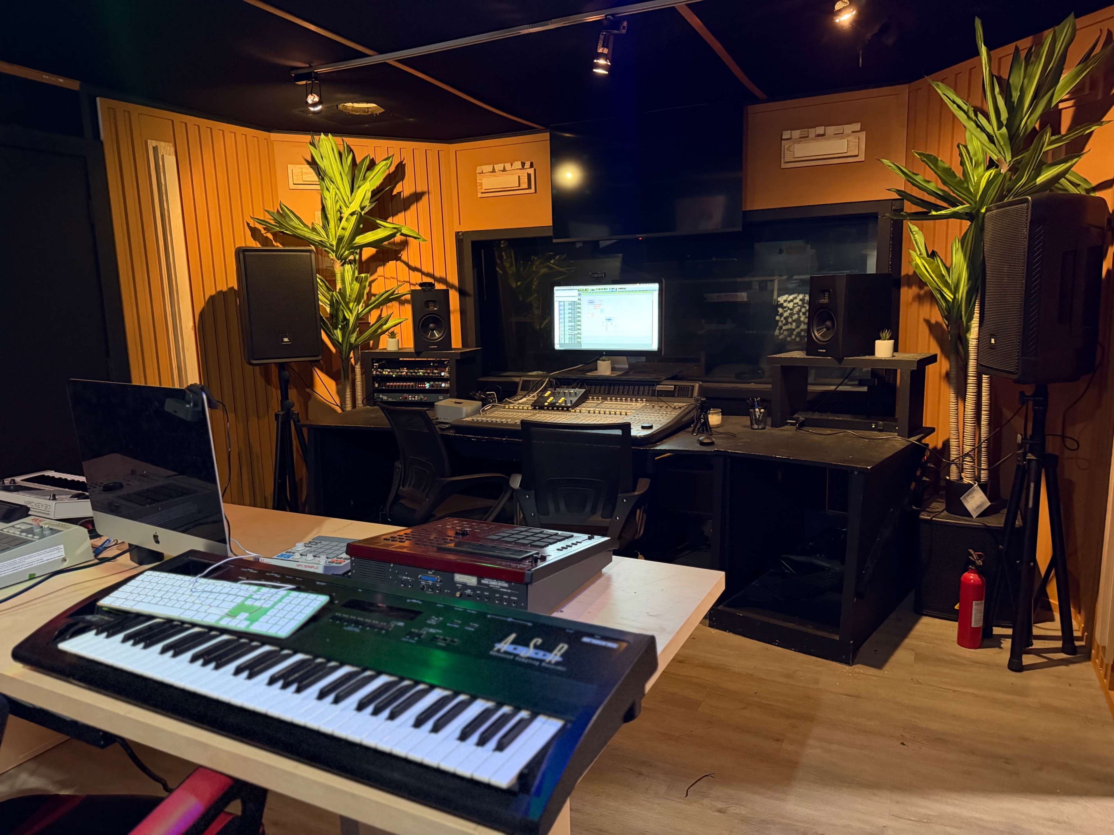 The image depicts a music studio equipped with a mixing console, monitors, speakers, and musical instruments, surrounded by wooden paneling and potted plants.