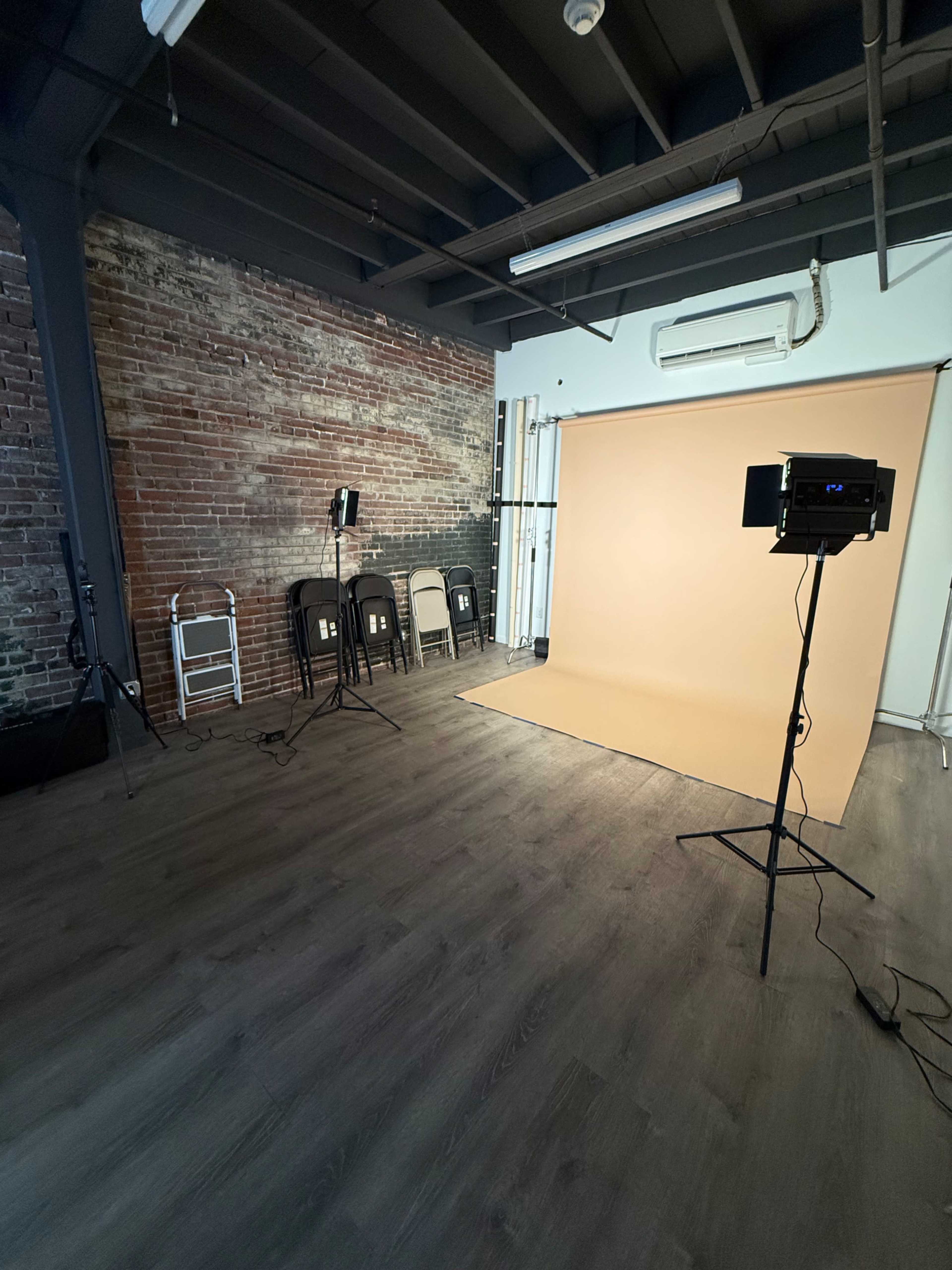 The image shows a photography studio with a backdrop, lighting equipment, and several black chairs arranged against a brick wall.