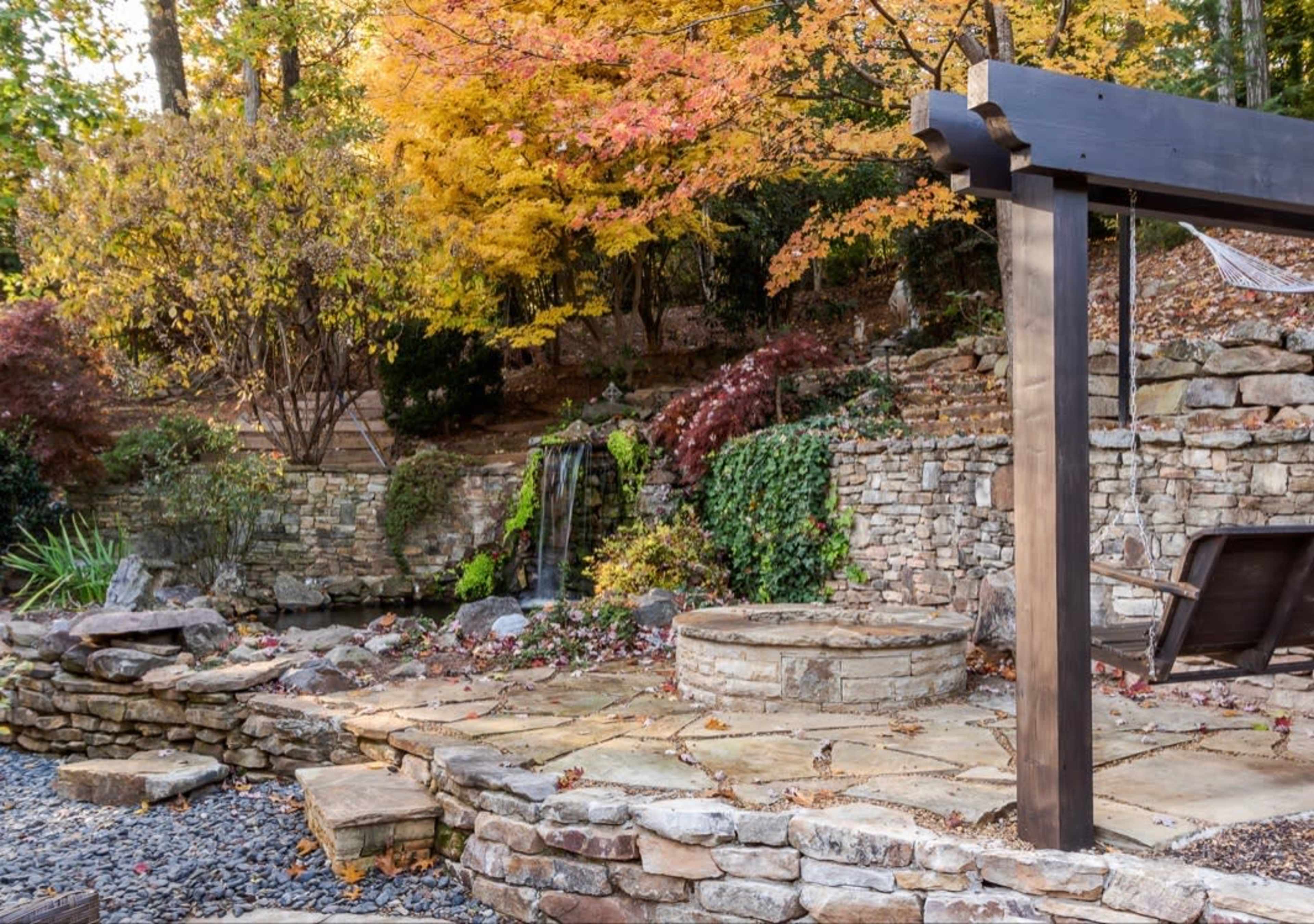 The image shows a landscaped outdoor area featuring a stone patio, a circular fire pit, and a small waterfall surrounded by autumn foliage.
