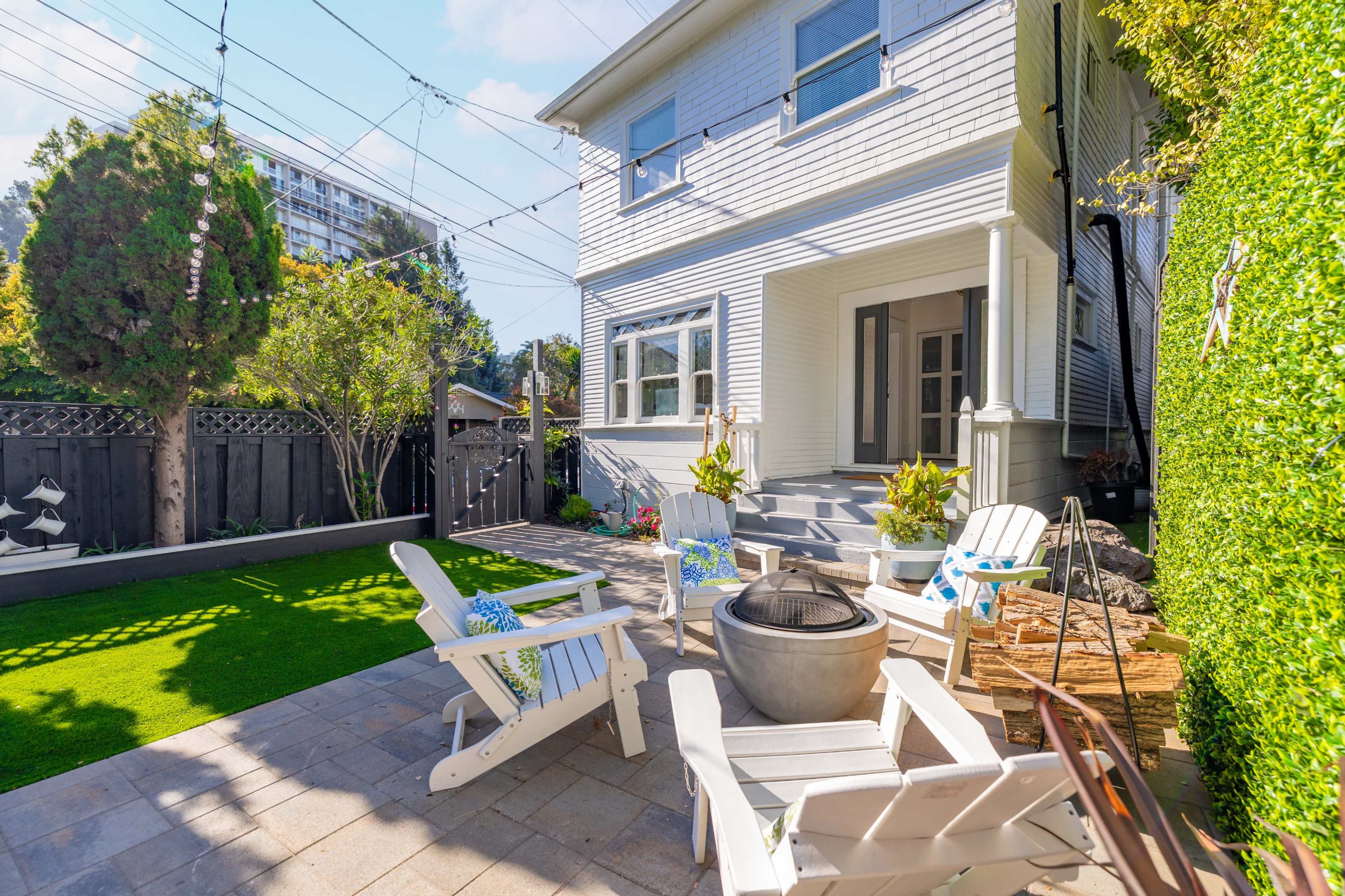A patio area features four white Adirondack chairs arranged around a metal fire pit, surrounded by greenery and a wooden fence.
