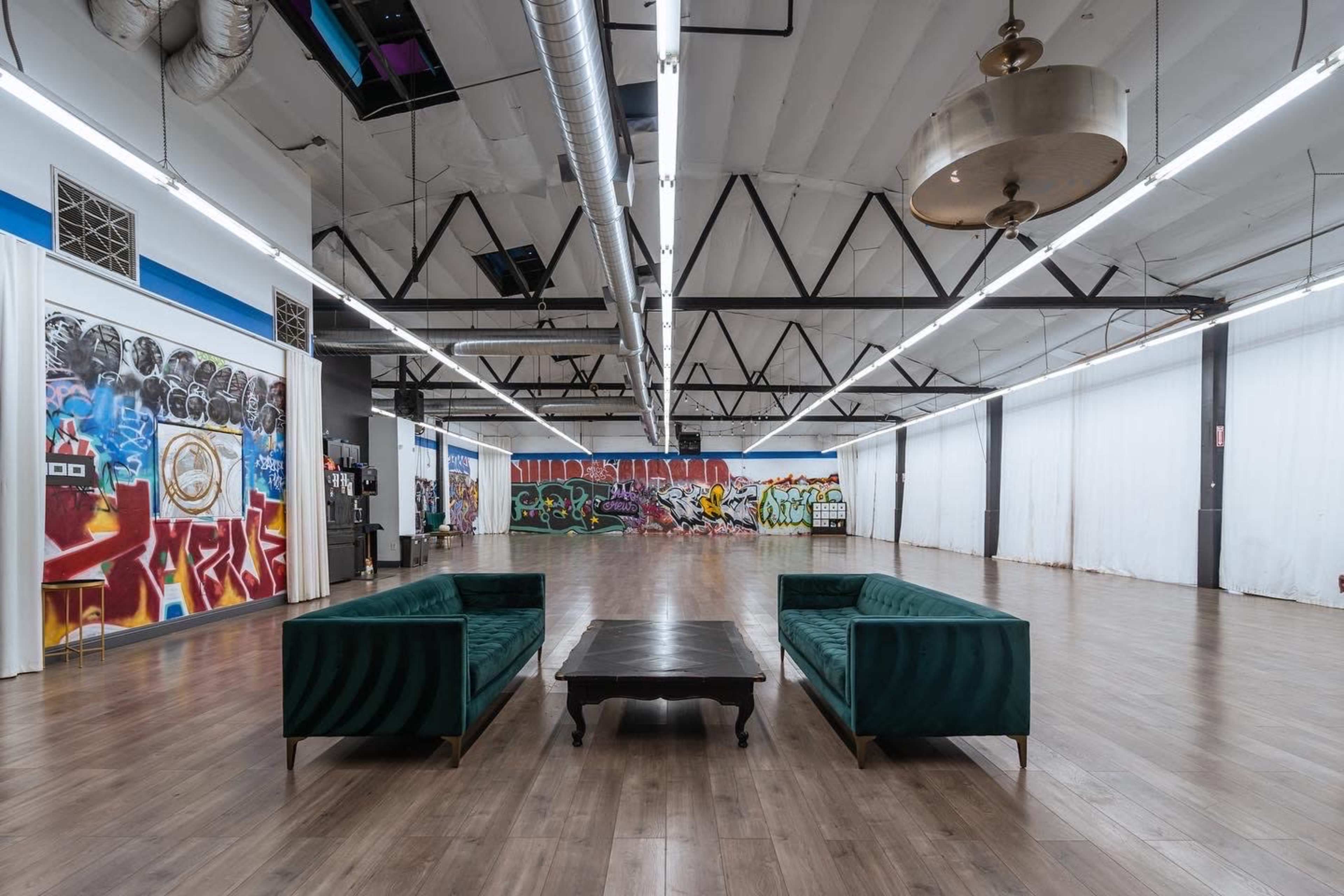 The image shows a spacious, modern room with polished wooden floors, featuring two green sofas and a central black table, along with graffiti artwork on the walls.