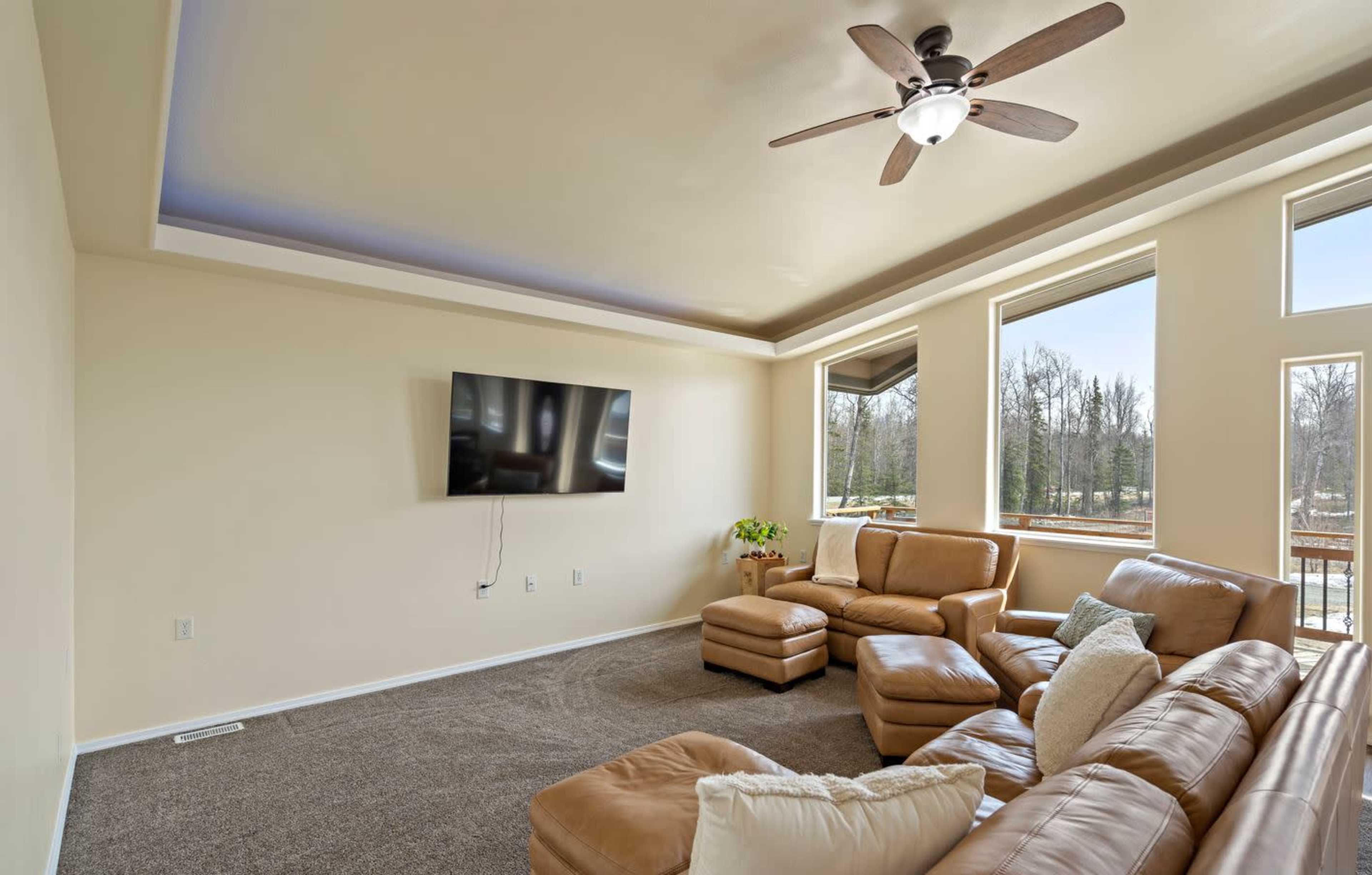 A spacious living room features a sectional sofa, a ceiling fan, and a wall-mounted television, with large windows providing natural light.