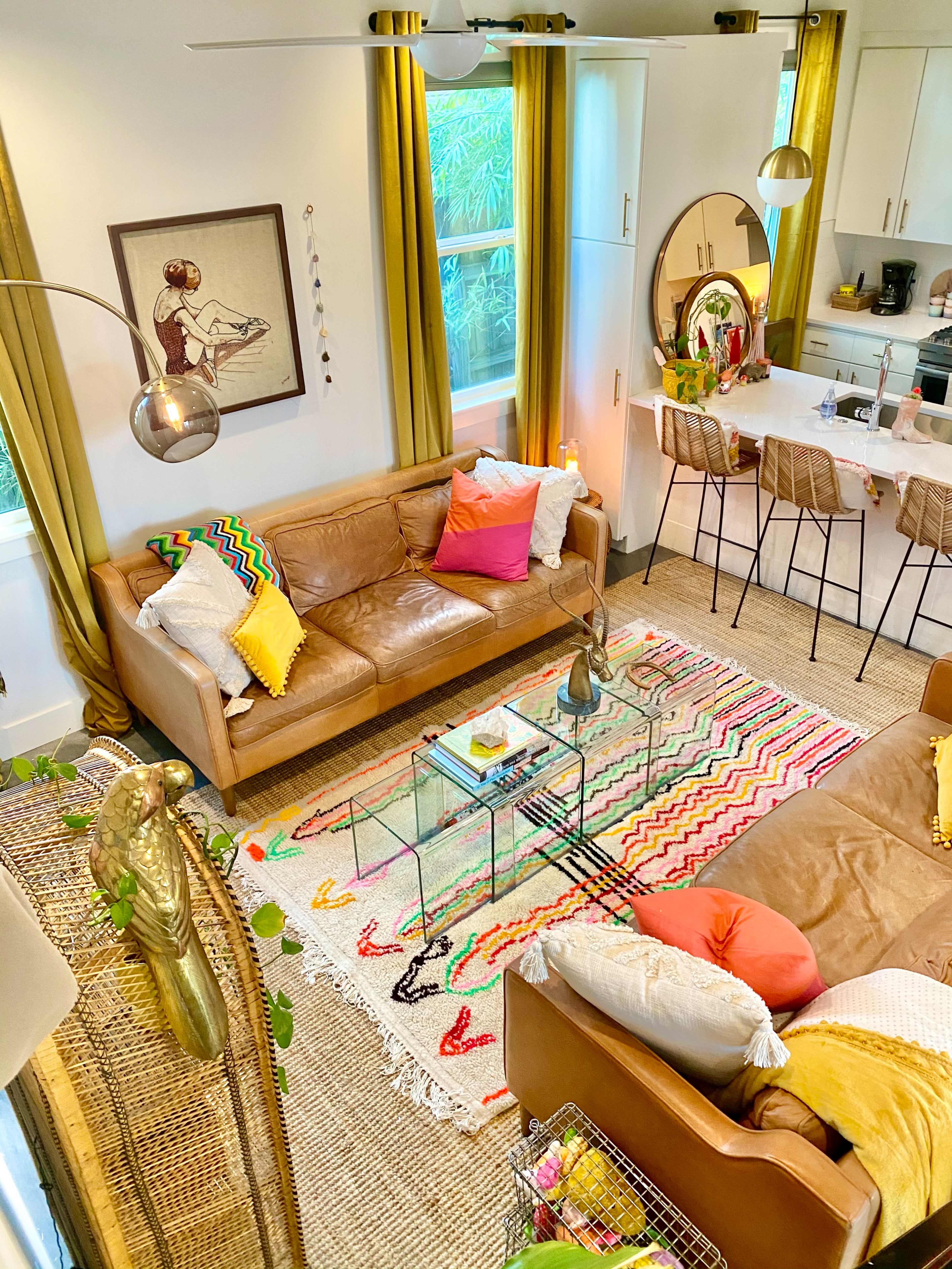 The image shows a brightly lit living room with a brown leather sofa, colorful throw pillows, a glass coffee table, and a kitchen area visible in the background.