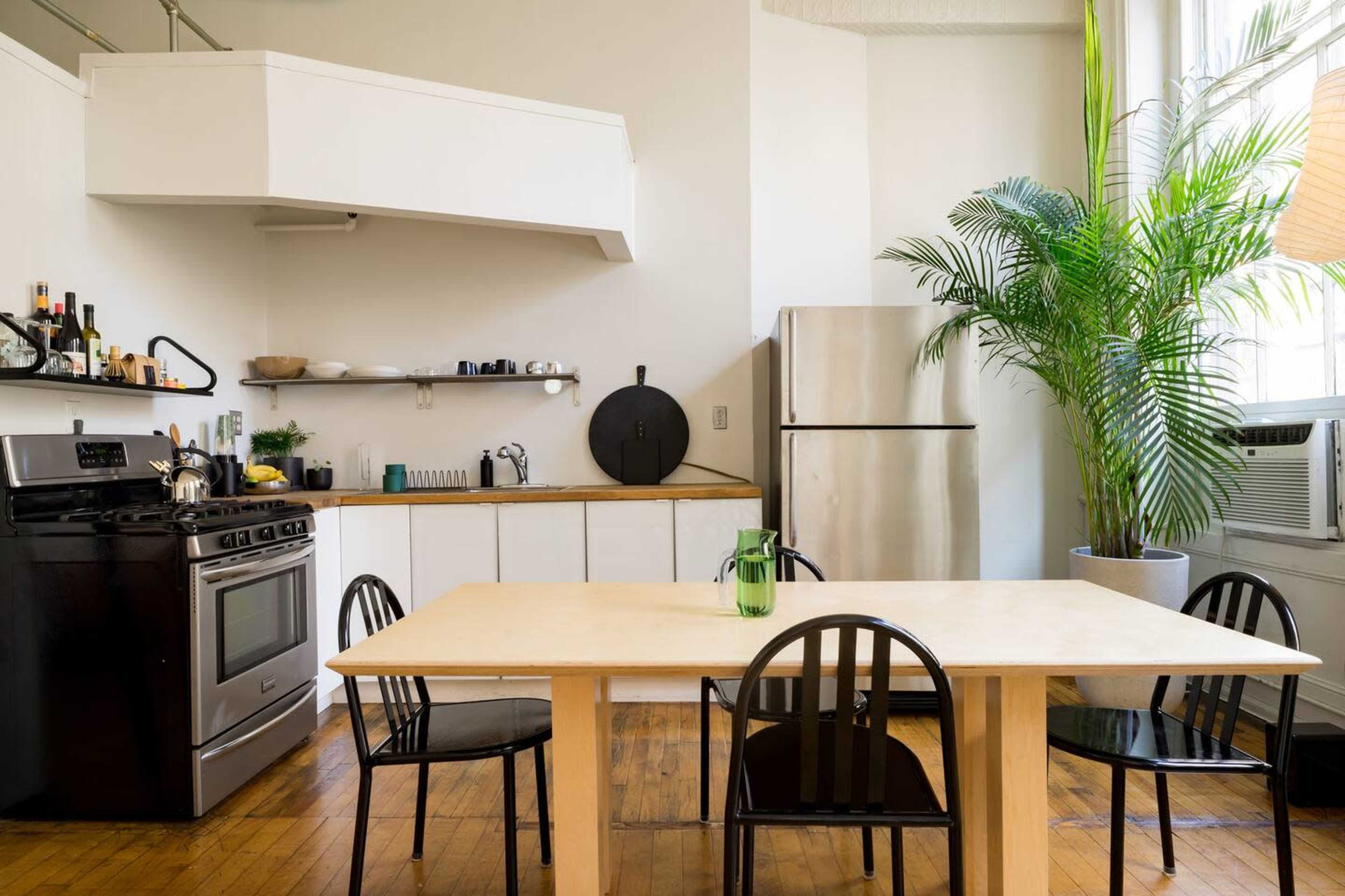 A modern kitchen features a black stove, stainless steel refrigerator, and a wooden table surrounded by black chairs, with plants adding a natural touch.