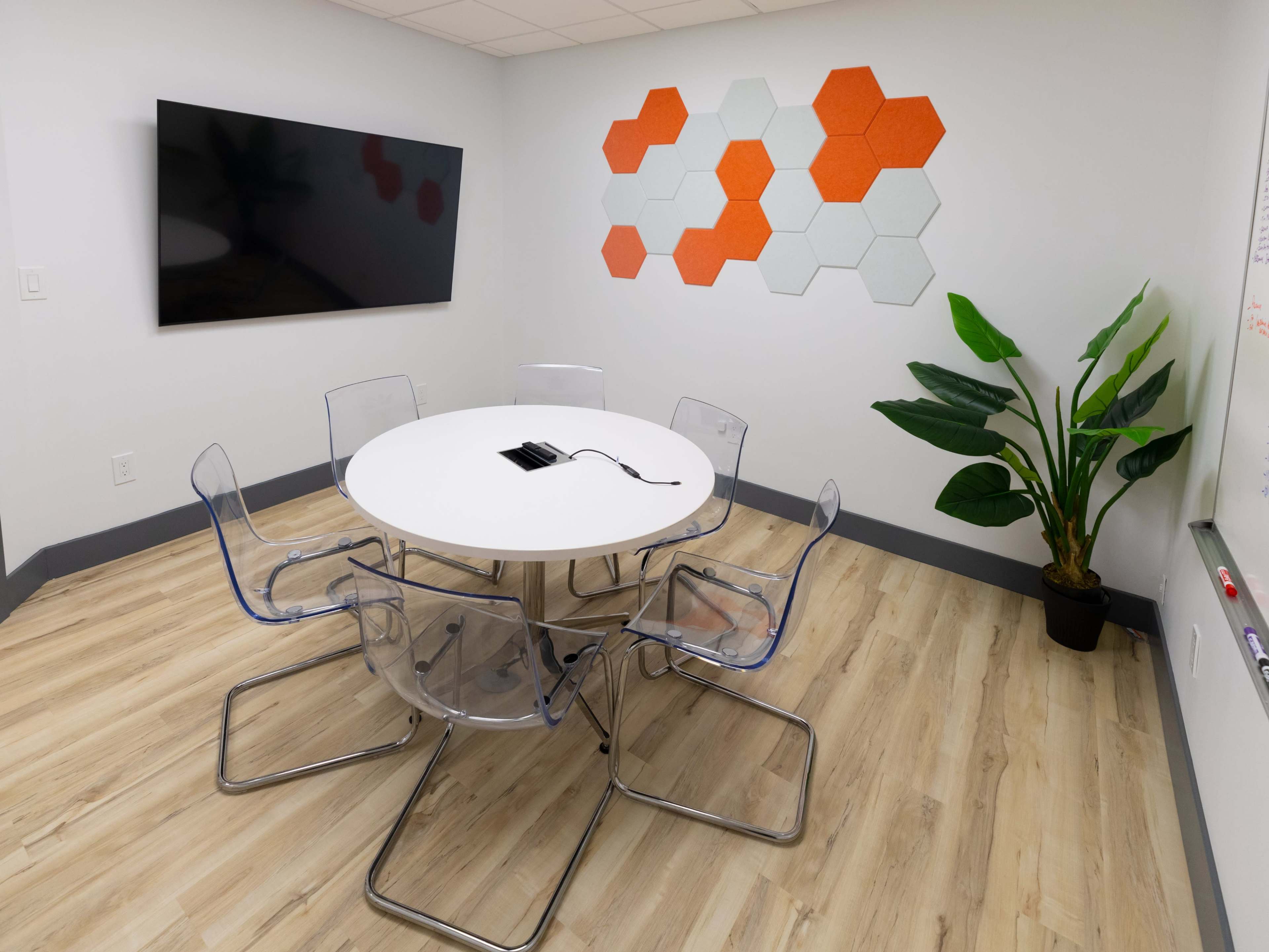 A conference room features a round table surrounded by transparent chairs, a large TV on the wall, and hexagonal orange wall accents.