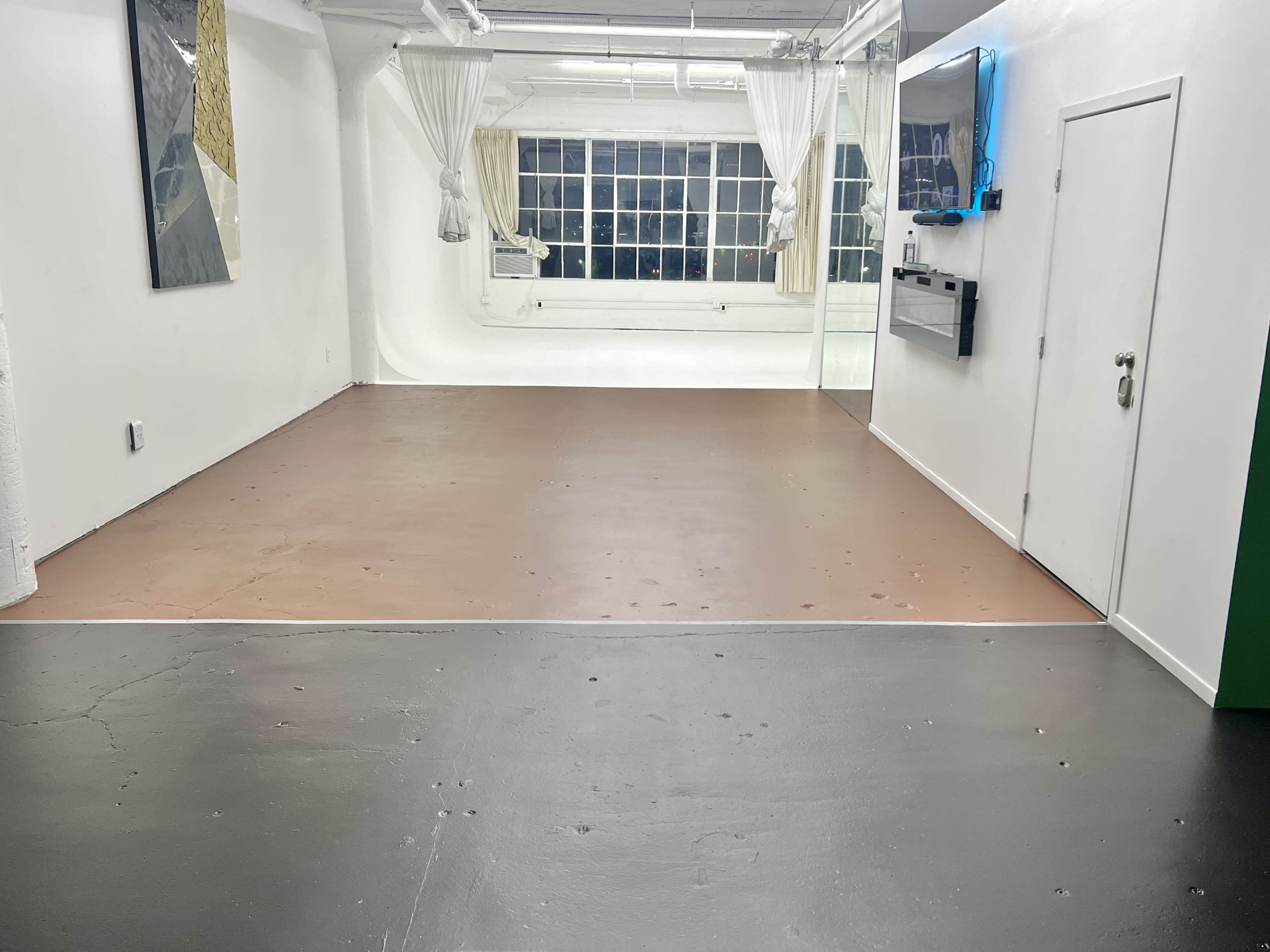 The image shows an empty room with a two-tone floor, featuring a brown area and a gray area, illuminated by natural light from large windows.