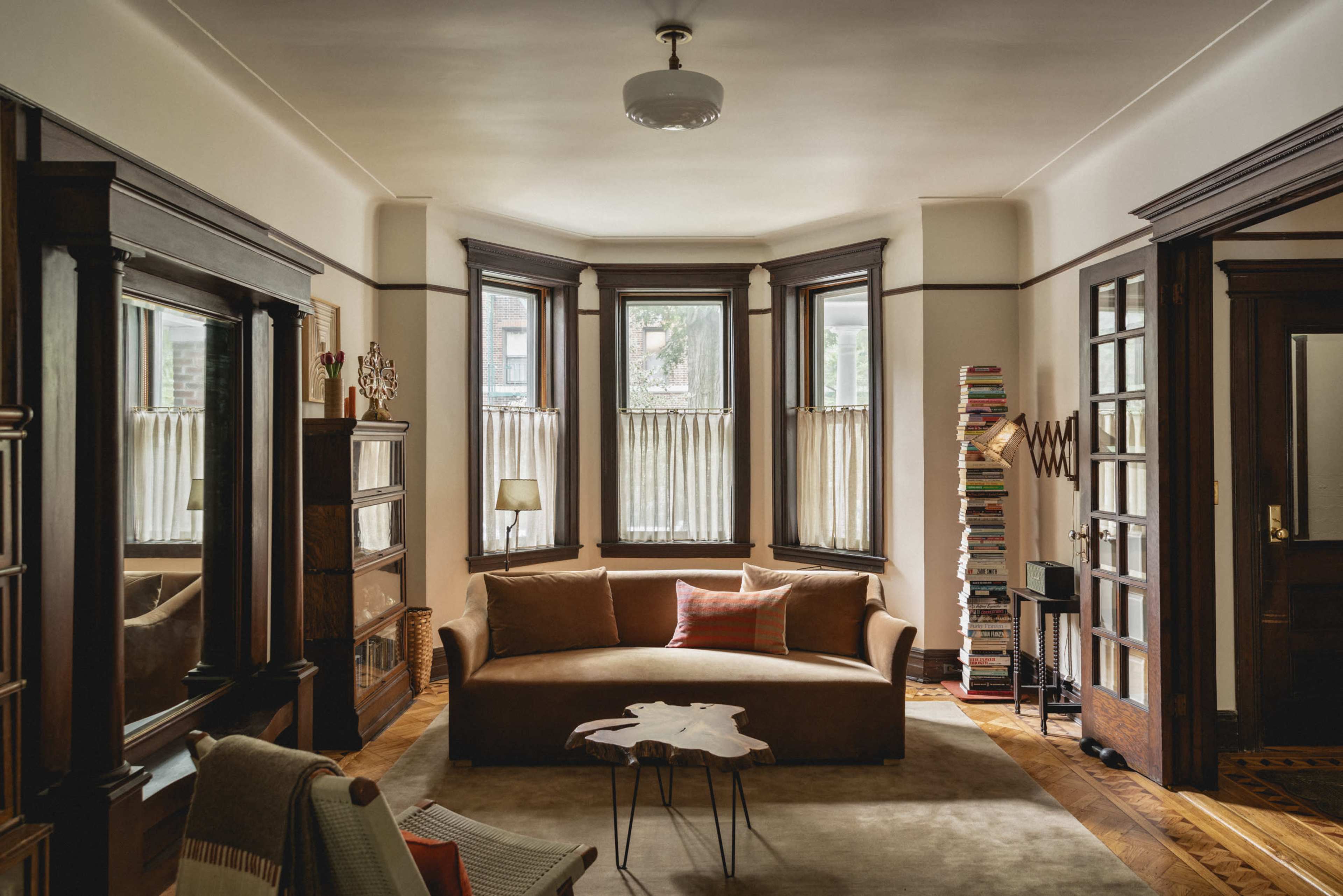 A living room with a brown sofa, an area rug, large windows with sheer curtains, and bookshelves filled with books.