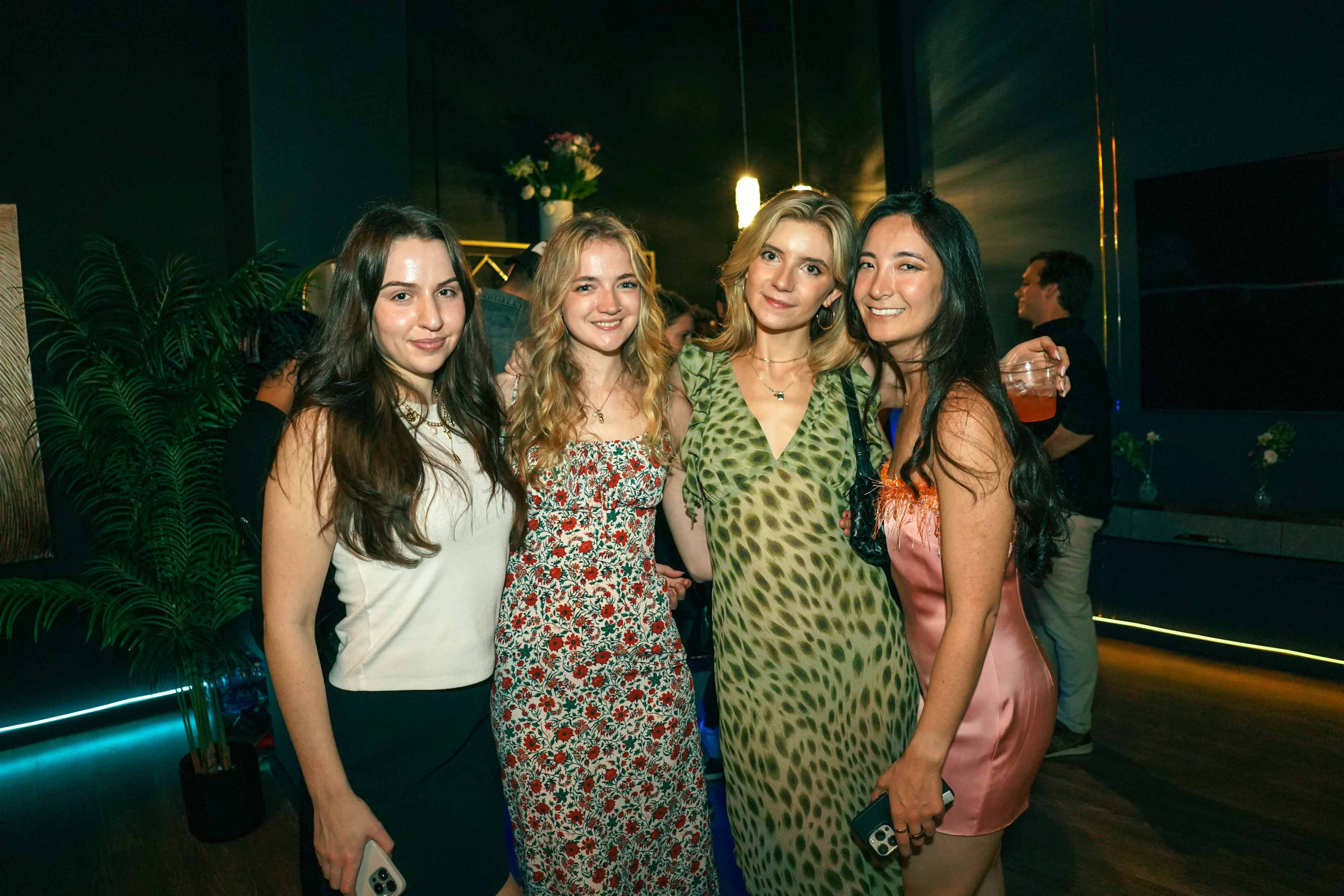 Four women pose together in a stylish interior with plants and decorative lighting in the background.